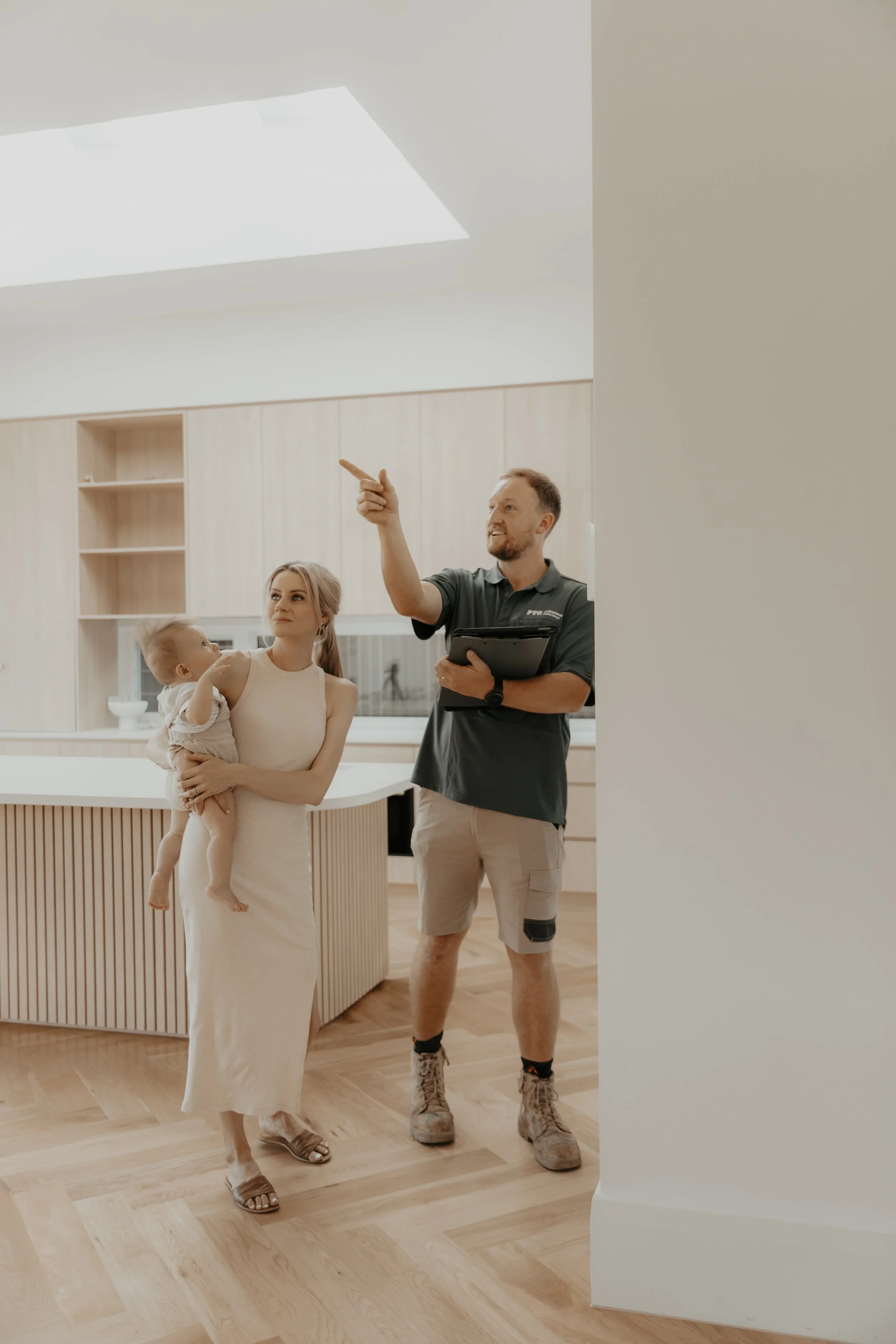 A woman holding a child stands in a modern kitchen with light wood cabinets, while a man in work attire points to the ceiling and holds a folder.