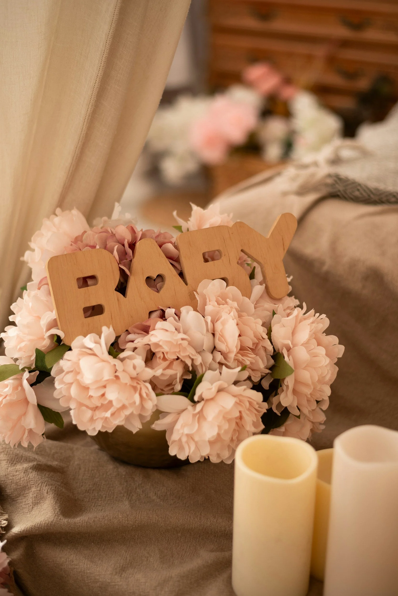 A floral arrangement of pink peonies with a wooden 'BABY' sign on top, placed on a beige tablecloth near cream-colored candles, in a softly lit room.