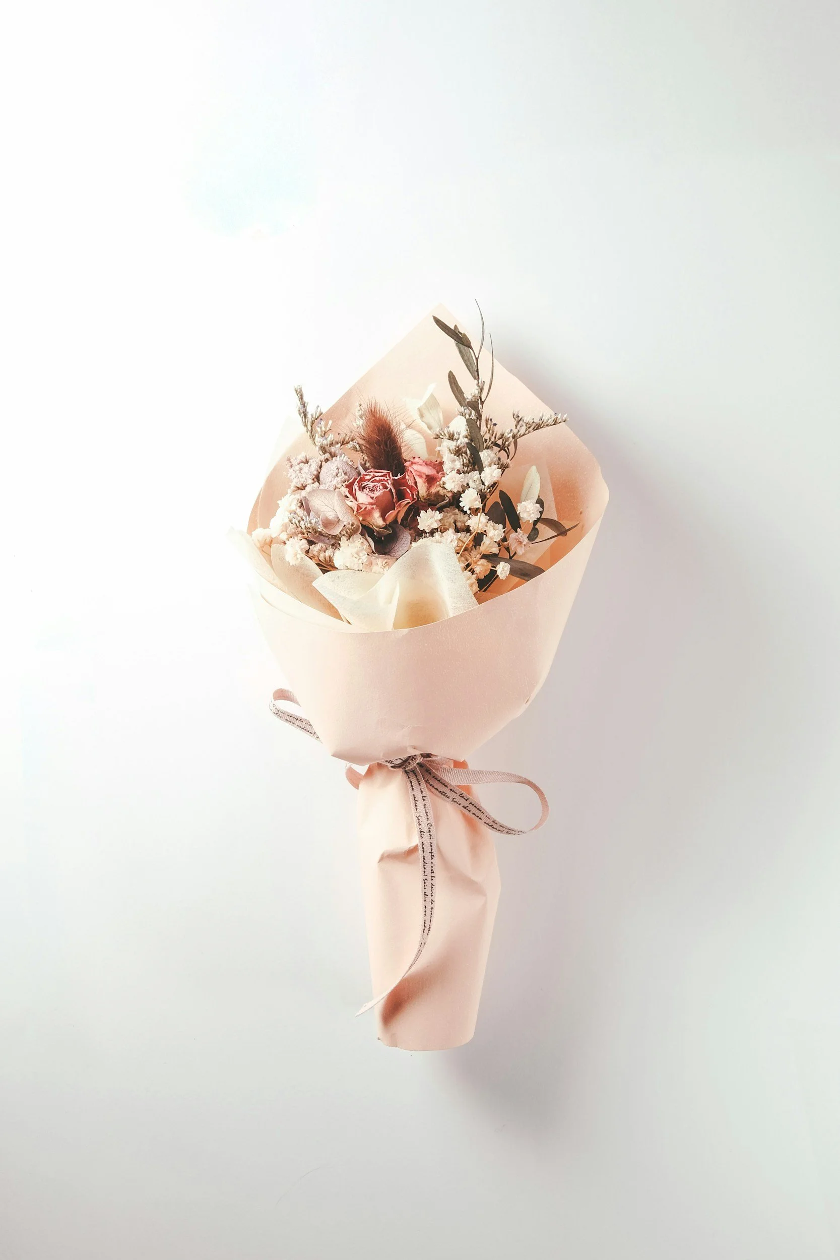 A pastel pink bouquet of dried and preserved flowers wrapped in pink paper with a ribbon tied around it. The bouquet is photographed against a white background.