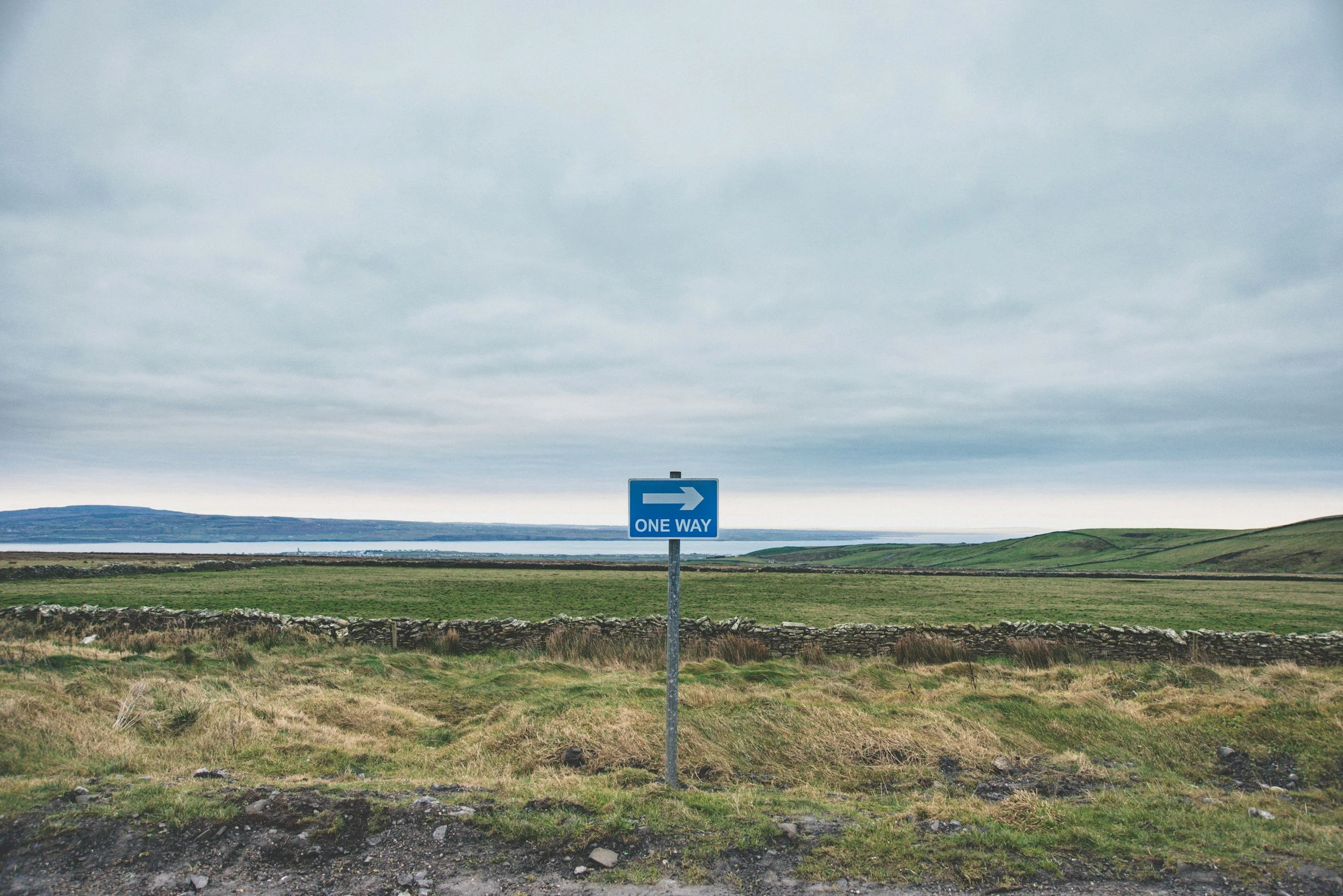 A blue one way traffic sign with an arrow pointing right, situated in a rural landscape with green fields, low stone walls, and hills in the background under a cloudy sky.