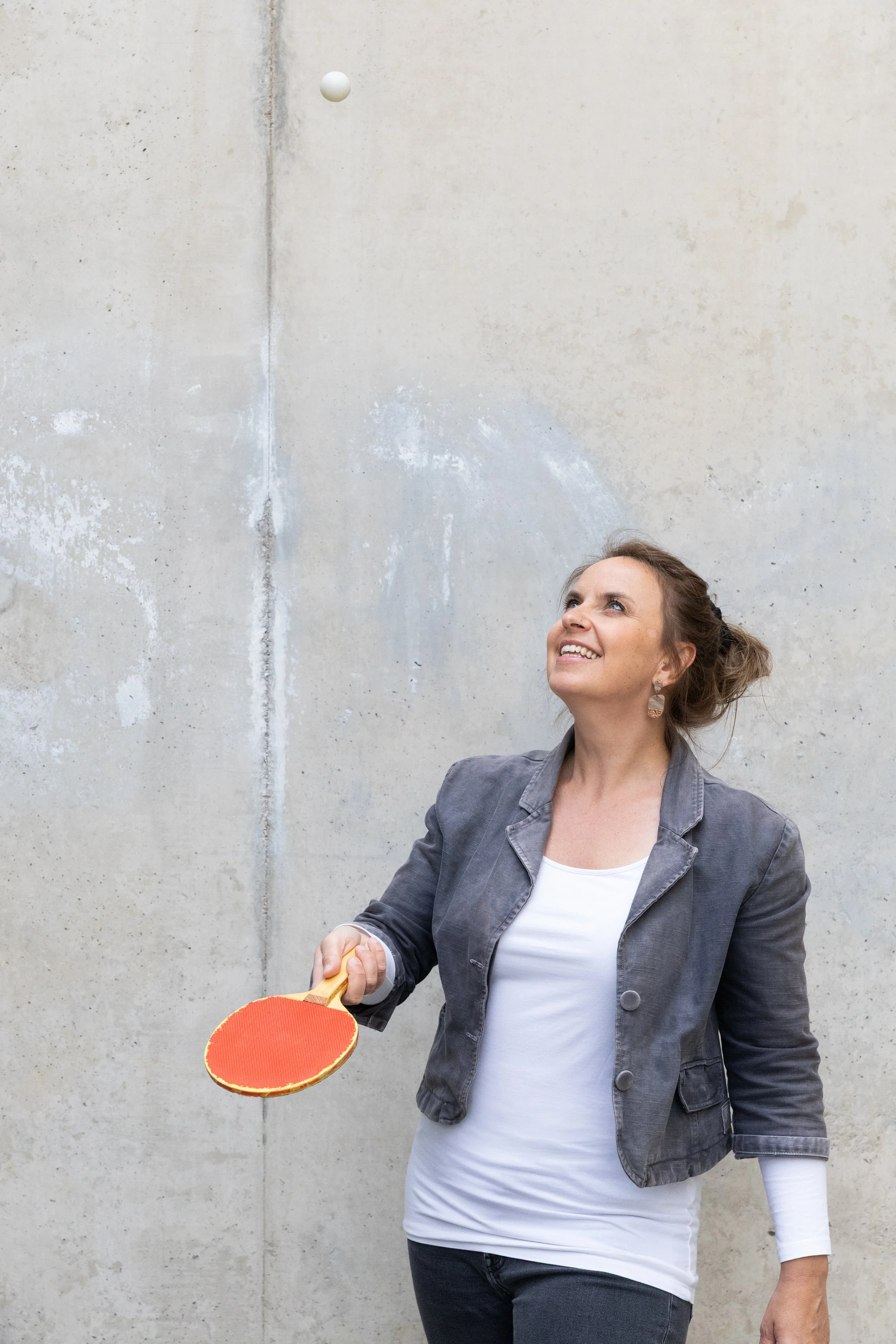 Eine Frau spielt Tischtennis gegen eine Wand. Sie hält einen Schläger und schaut lachend nach oben. Es sieht so aus, als würde die weiße Tischtennisball fliegen.