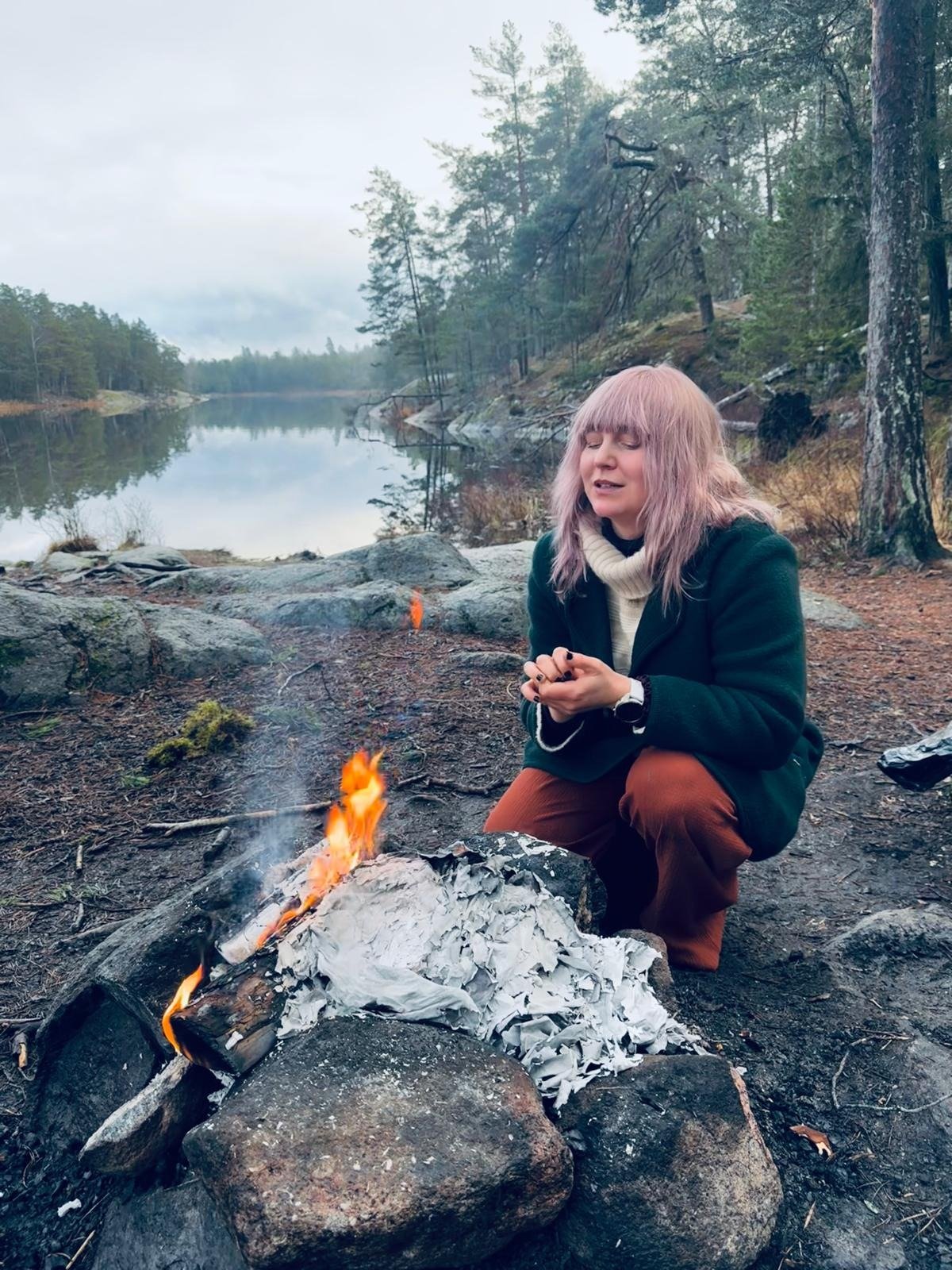 Woman kneeling by a campfire near a forest lake during a ritual.