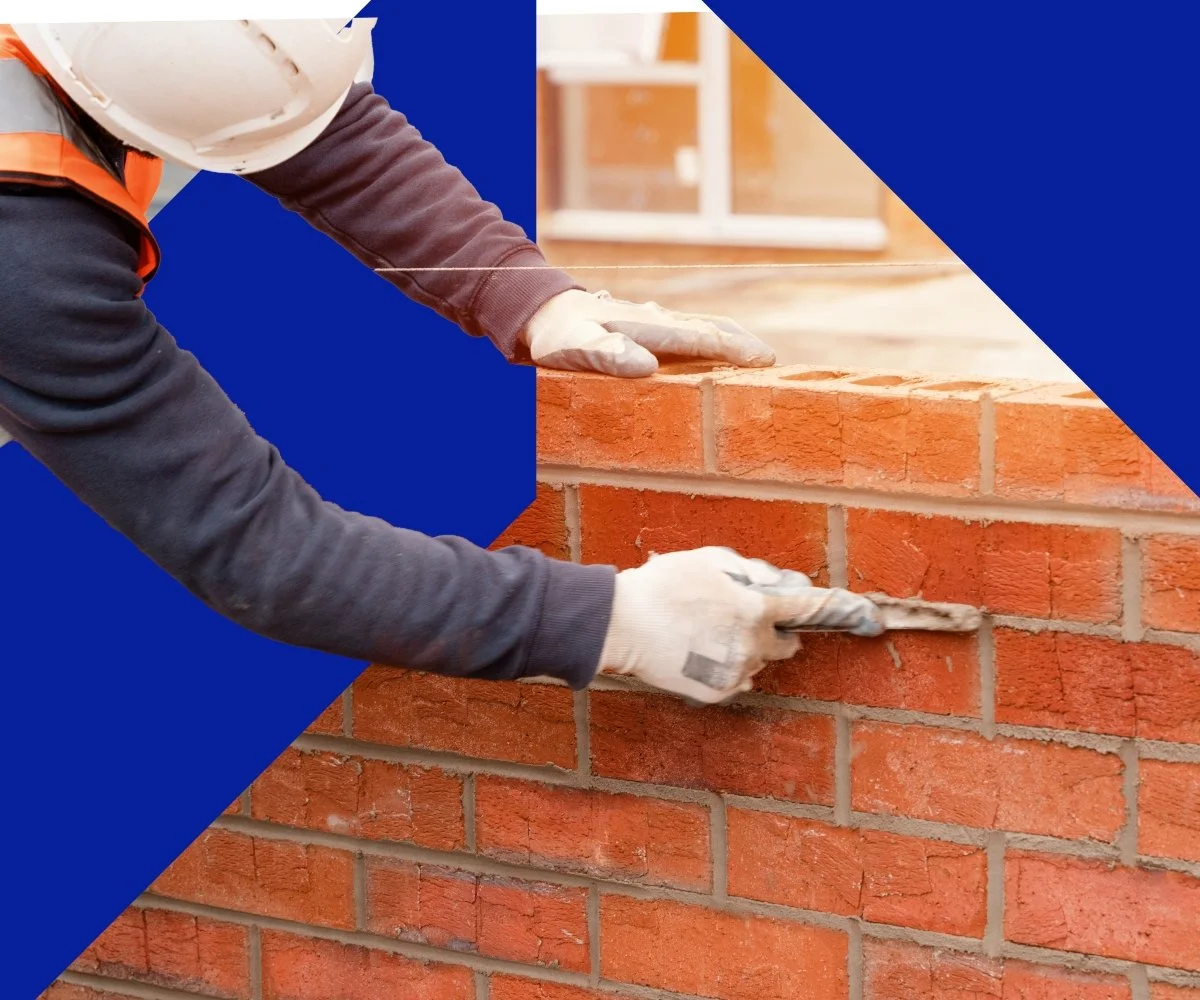 Construction worker laying red bricks to build a wall, wearing gloves, a hard hat, and safety attire.