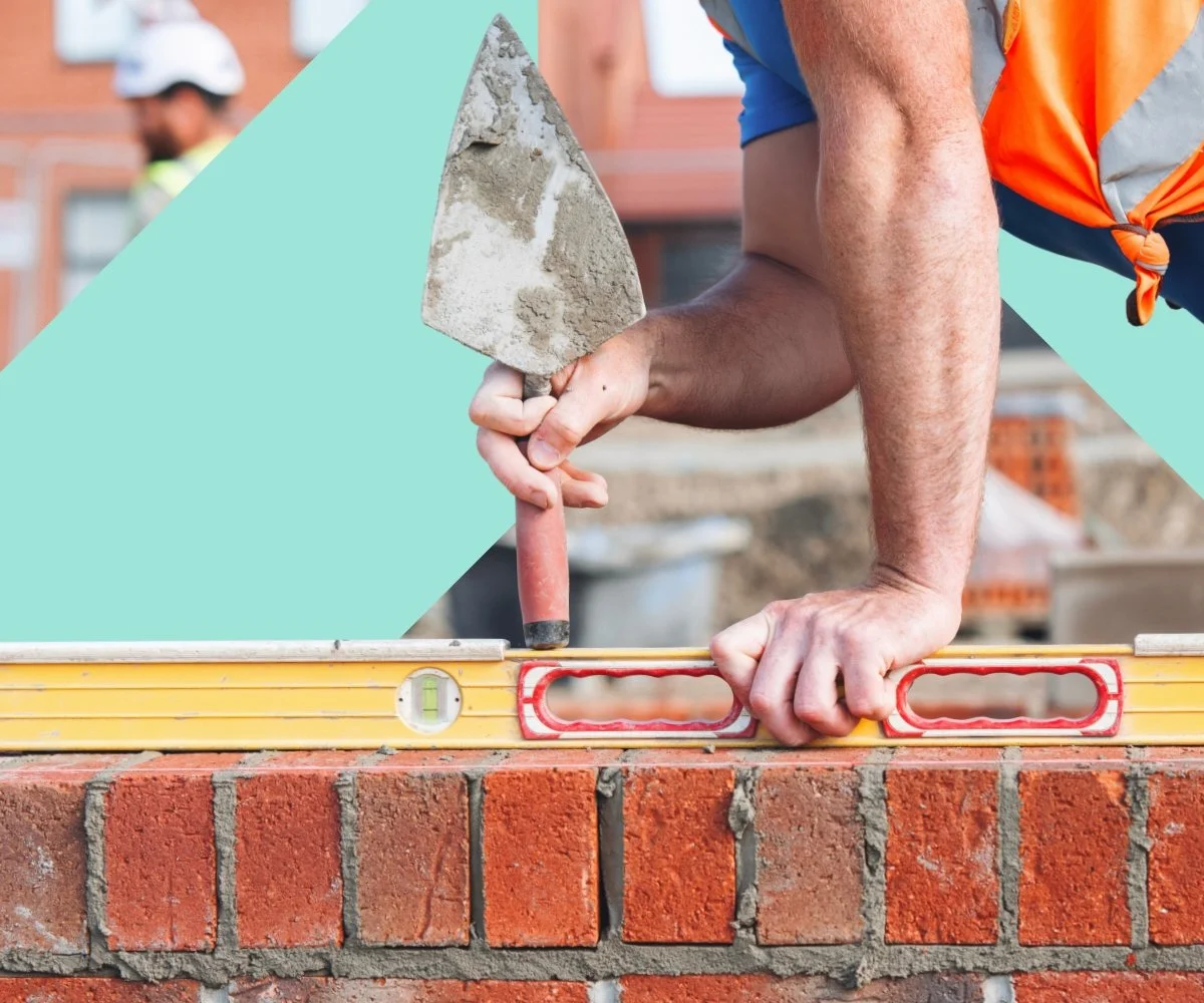 A construction worker laying bricks uses a trowel and level on a brick wall at a building site.