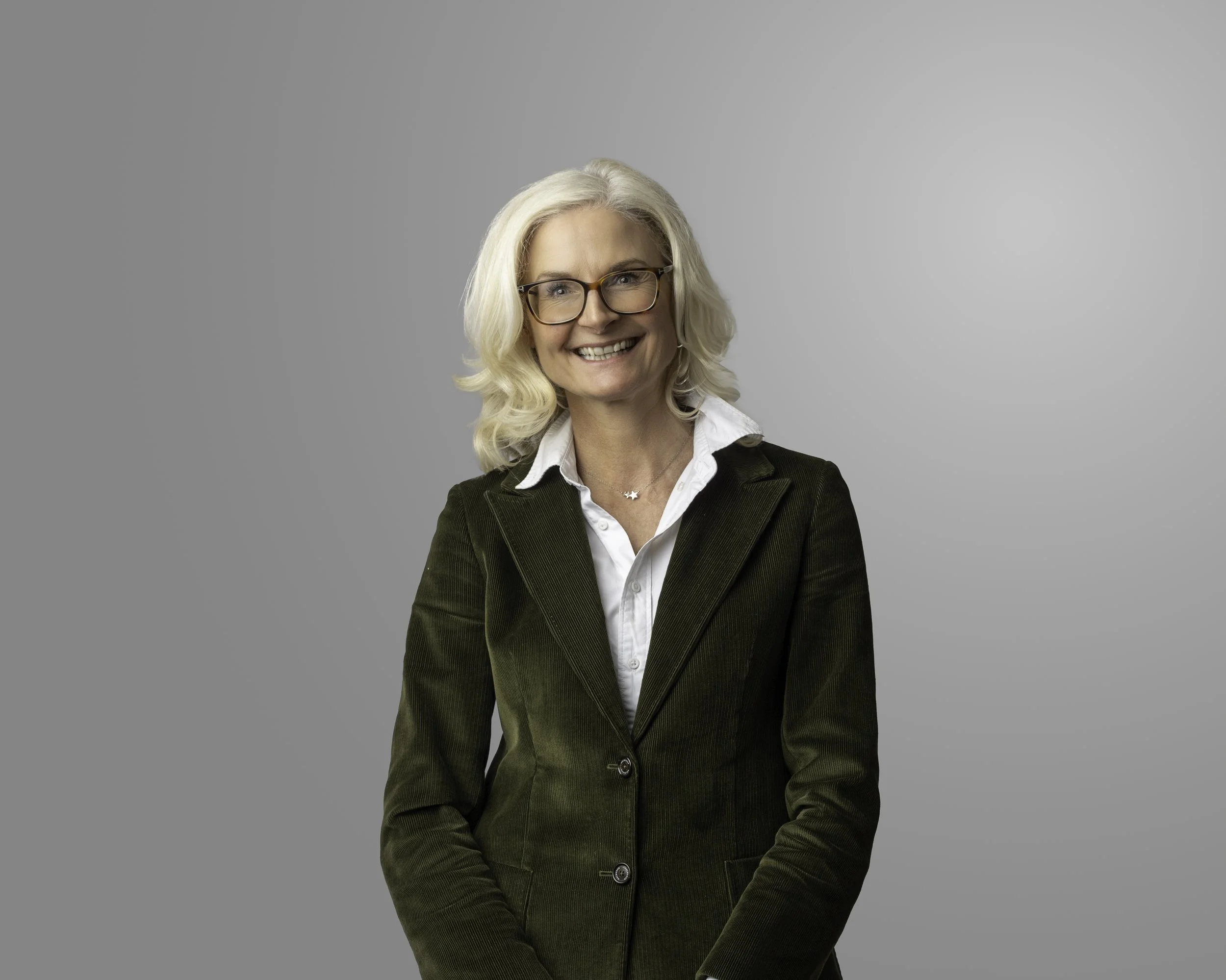 Smiling middle-aged woman with blonde hair, glasses, white shirt, navy sweater, green jacket, and bird necklace standing outside in front of a brick wall and window.
