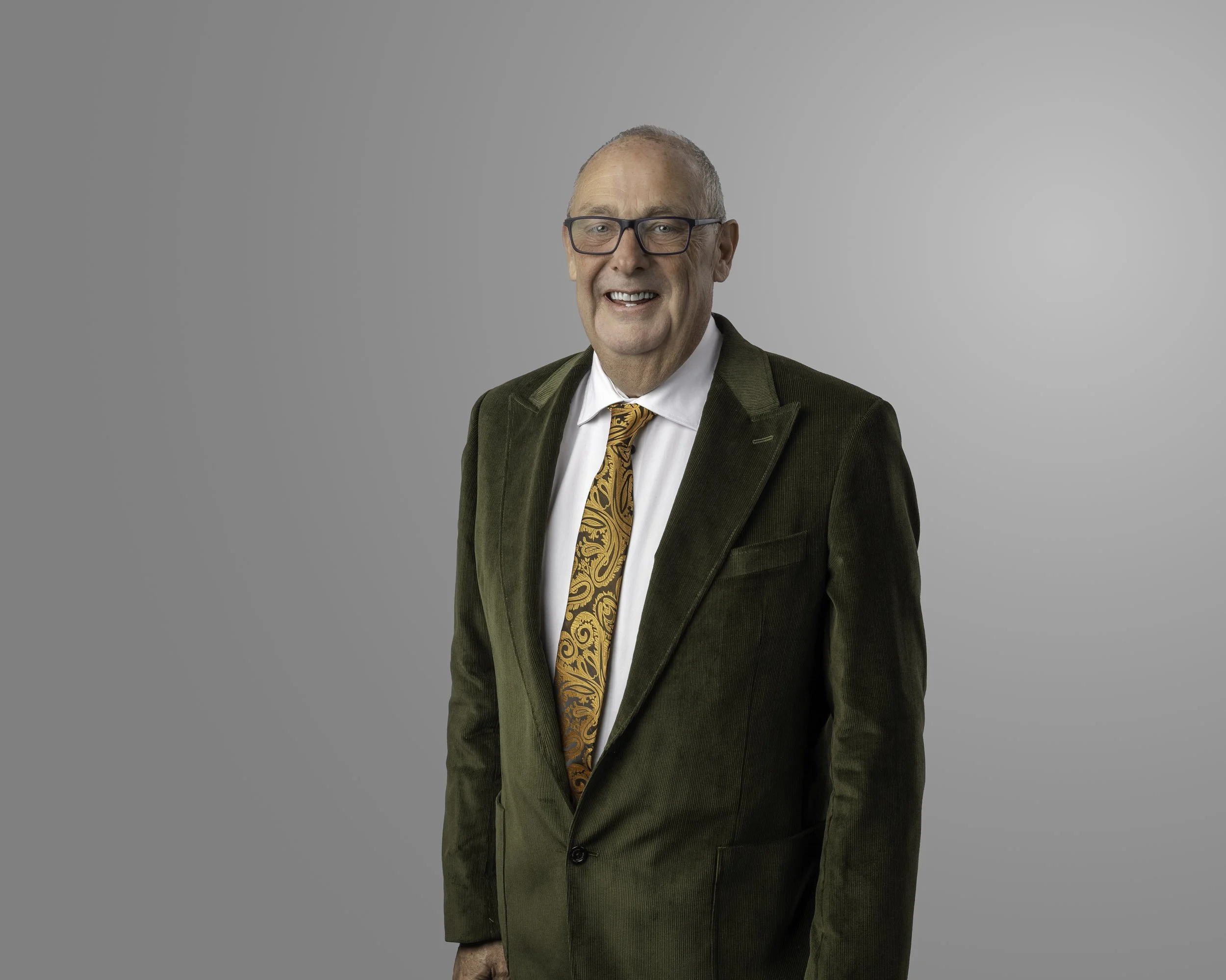 A smiling elderly man with a white beard and short hair, dressed in a suit and tie, standing outside near a brick wall with a window in the background.