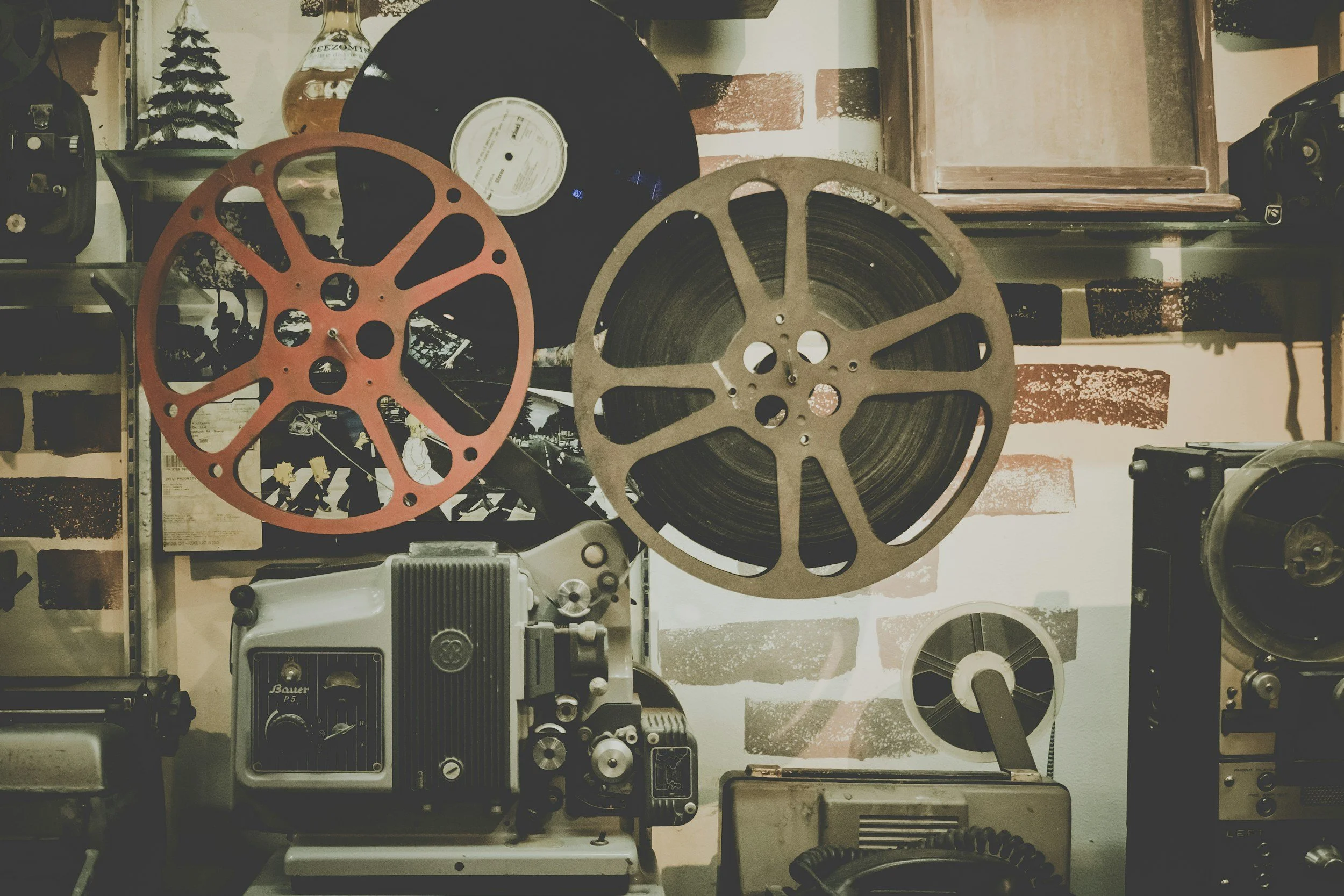 A collection of film reels, a vintage film projector, and other film equipment on shelves against a wall with a brick pattern.