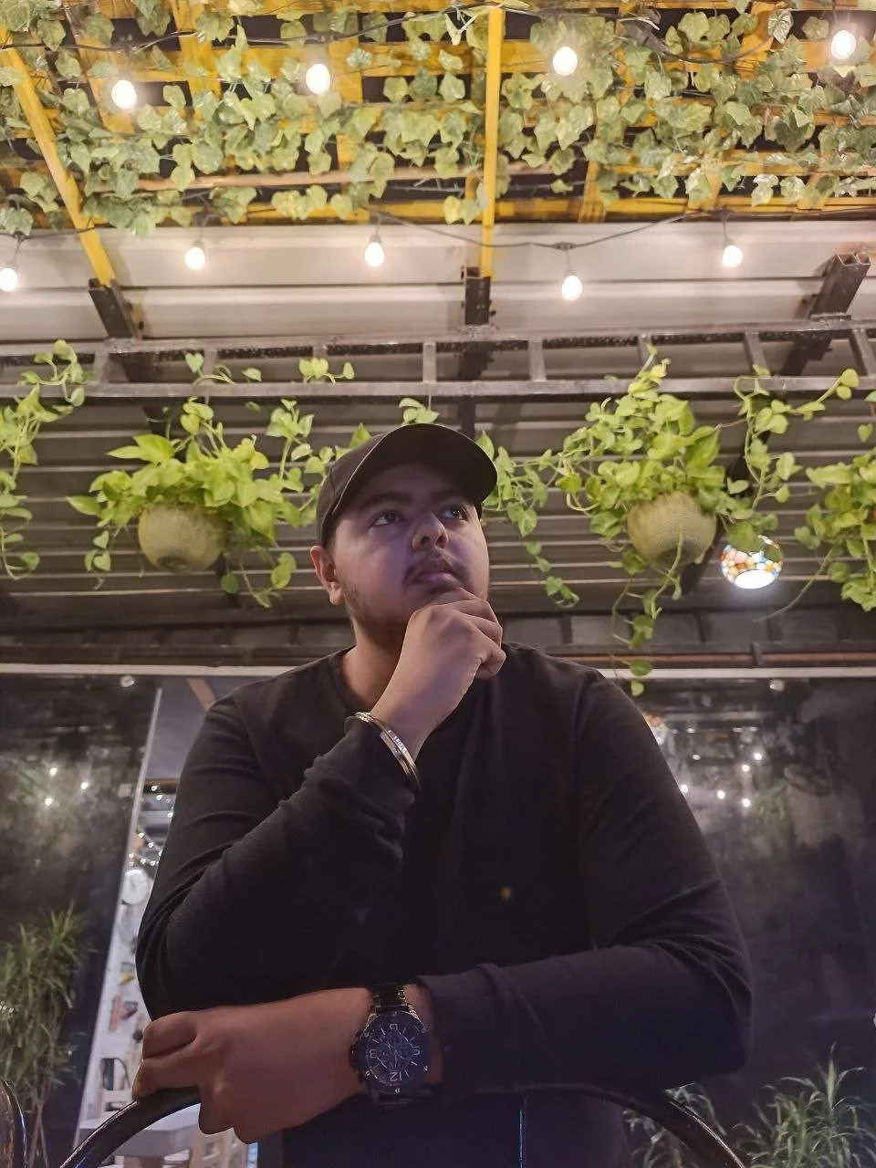 A young man wearing a black shirt and a black cap sitting at a table in a restaurant or cafe, with his hand resting on his chin, looking thoughtful. The background features hanging green plants and decorative hanging lights.