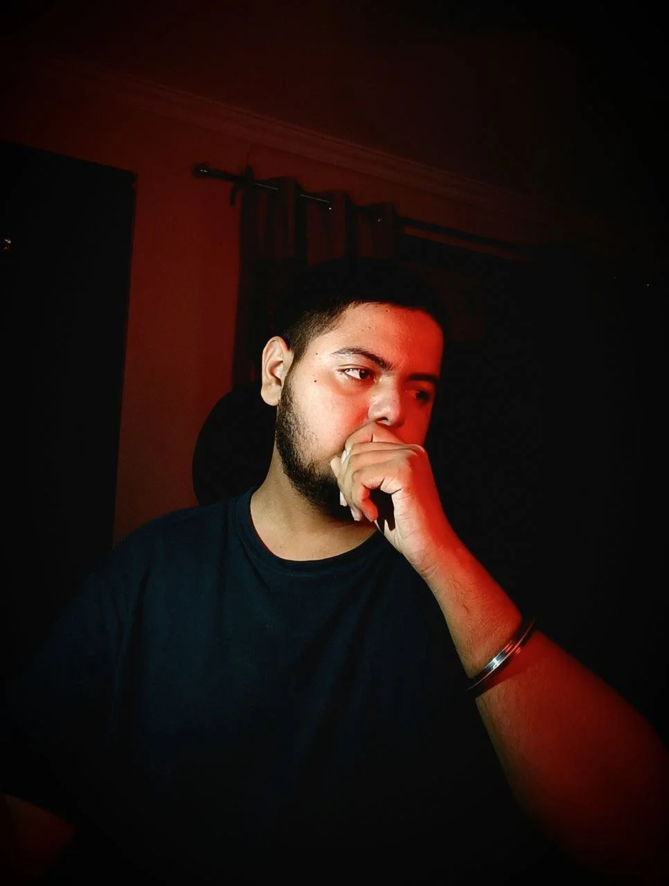 A young man with a beard and short hair sits in a dark room with red lighting, resting his chin on his hand, wearing a black t-shirt and a wristband.