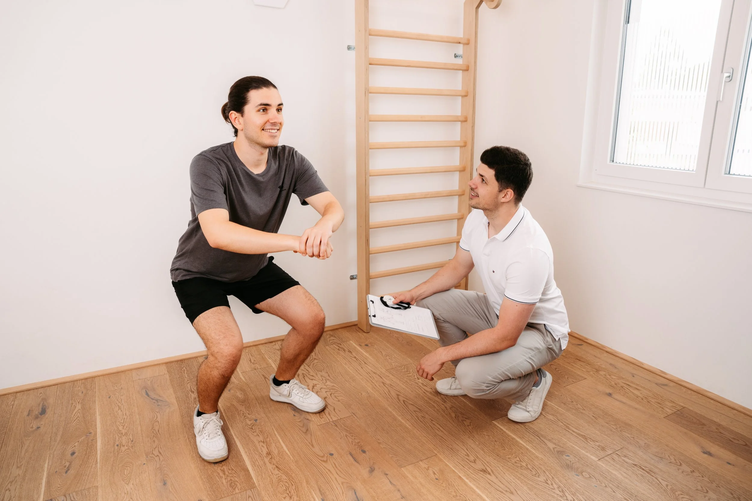 Zwei Männer beim Physiotherapie-Check in einem Raum mit Holzfußboden und Turnwand, ein Mann macht Squats, der andere hält eine Checkliste.