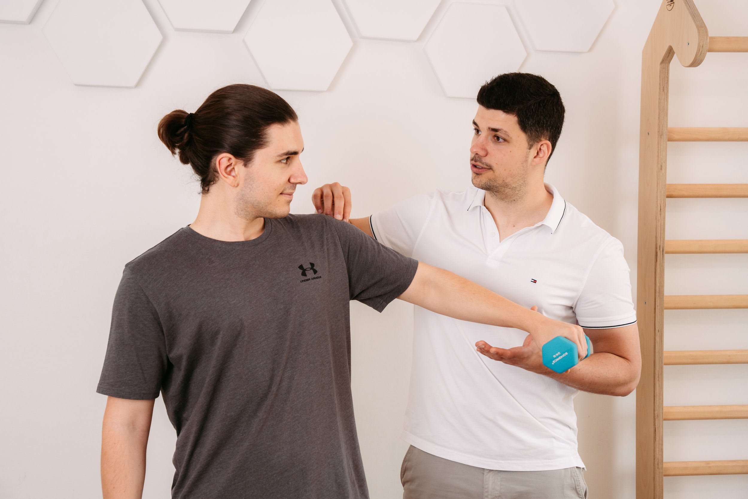 Ein Personaltrainer macht für einen Mann ein Dehnungs- und Dehnübung im Fitnessstudio oder Physiotherapie. Der Trainer steht neben dem Mann, der eine graue Sport-T-Shirt trägt, und hält seine Schulter, während er mit einer blauen Hantel in der Hand die Arme streckt. Im Hintergrund ist eine Holzstange und eine Wand mit weißen, geometrischen Dekorationen.
