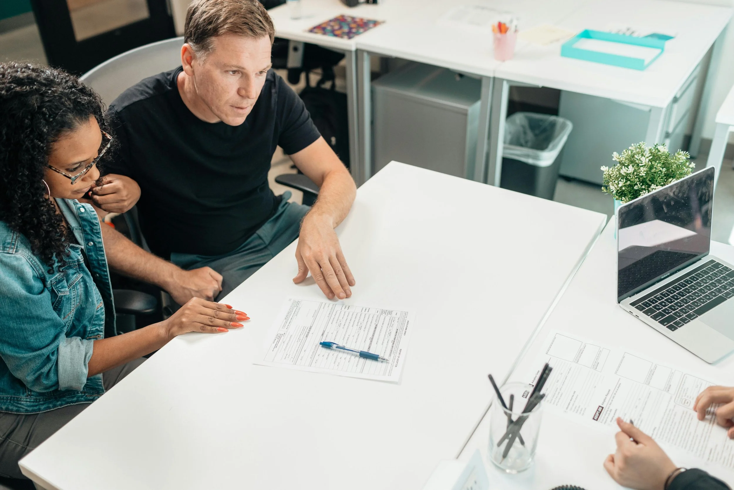 Two people, a woman with glasses and curly hair and a man with short hair, sit at a white table discussing documents. There is a laptop, a potted plant, a pen, and other office supplies around them in an office setting.
