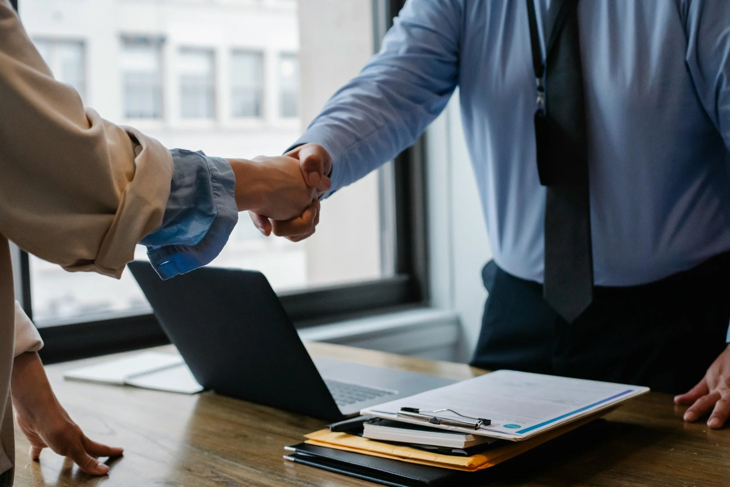 Two people shaking hands over a desk with a laptop, documents, and folders in an office.