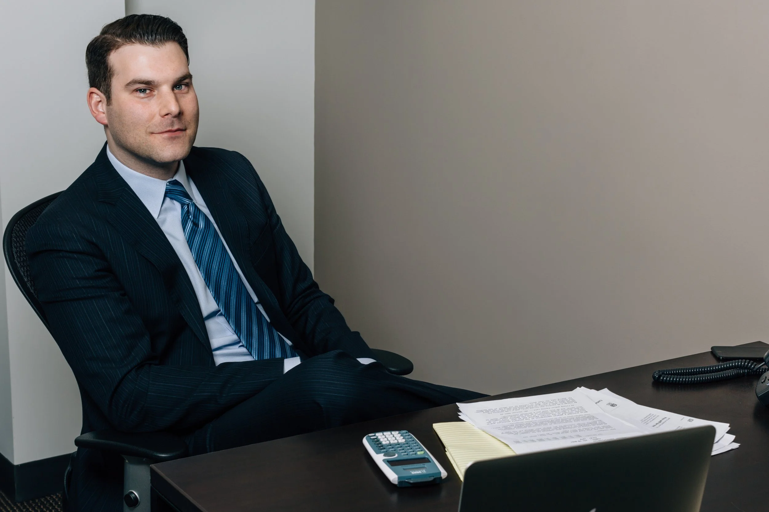 A man in a dark pinstripe business suit with a light blue shirt and a striped blue tie sits at a desk in an office, looking at the camera. The desk has papers, a yellow notepad, a calculator, and a landline phone.