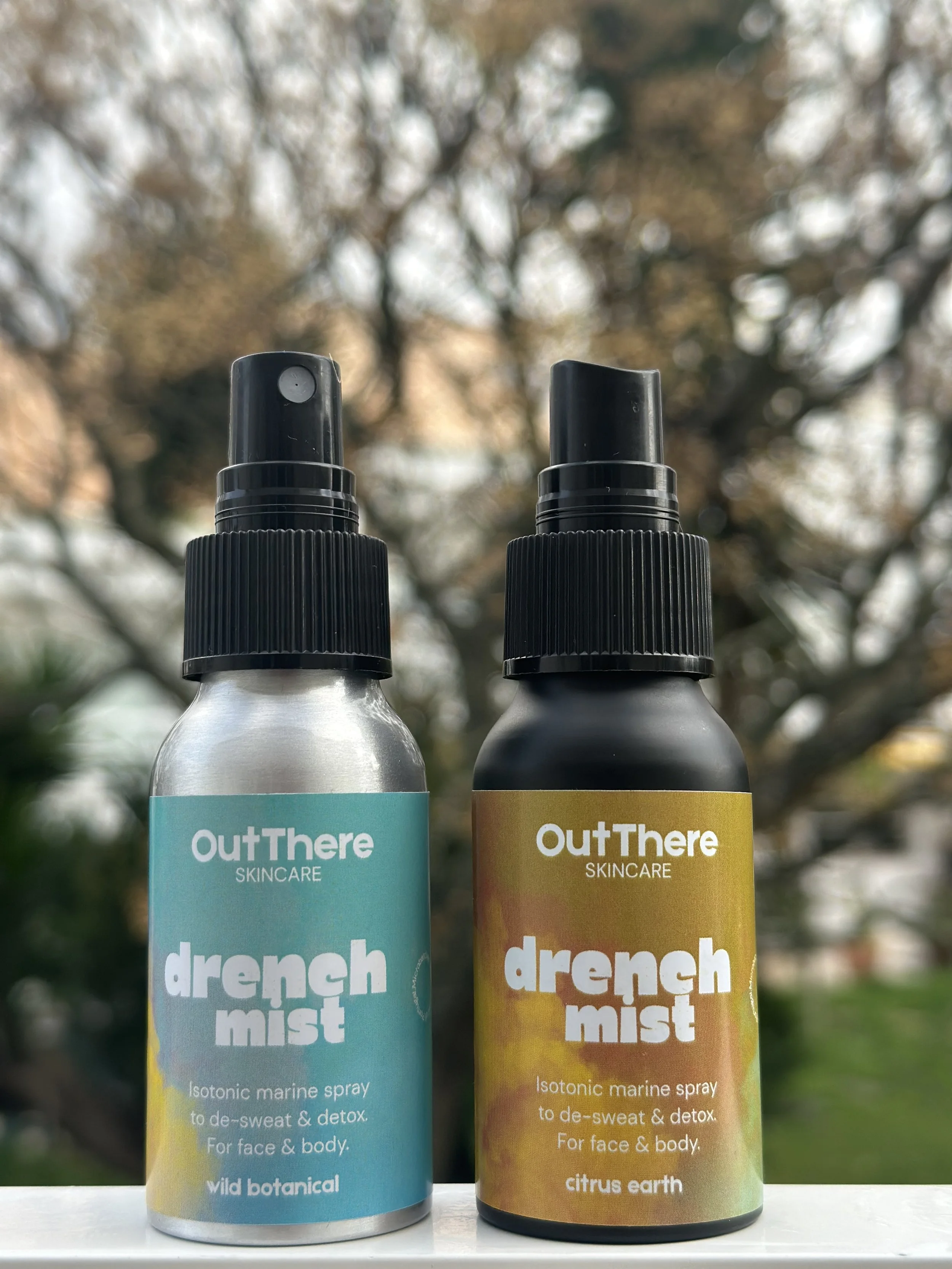 Two bottles of OutThere Skincare Drench Mist, one with a blue label and the other with a yellow label, set outdoors with blurred trees in the background.