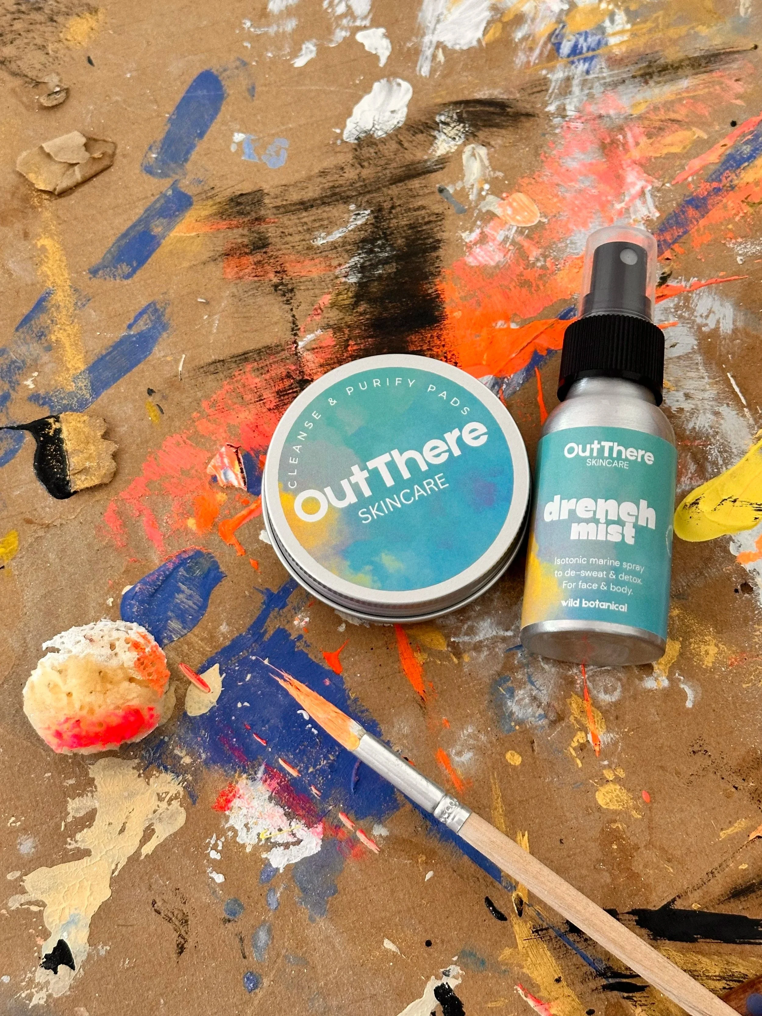 Skincare products with OutThere branding, including a jar of cleanse and purify pads and a spray bottle of Drench Mist, placed on a paint-splattered wooden surface.