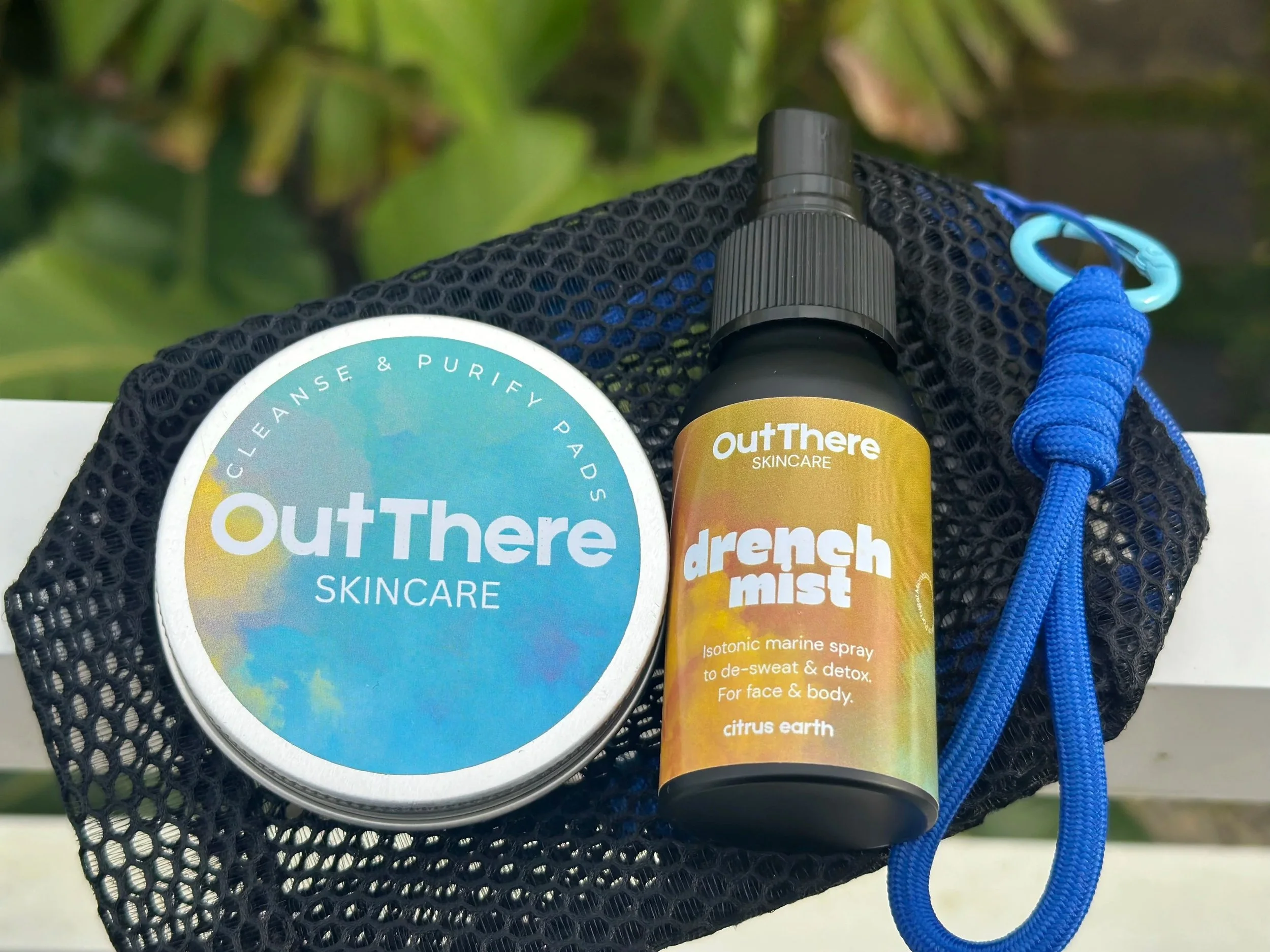 Skincare products, OutThere brand, cortical and detox, and marine spray on black mesh bag outside, with lush green foliage in background.