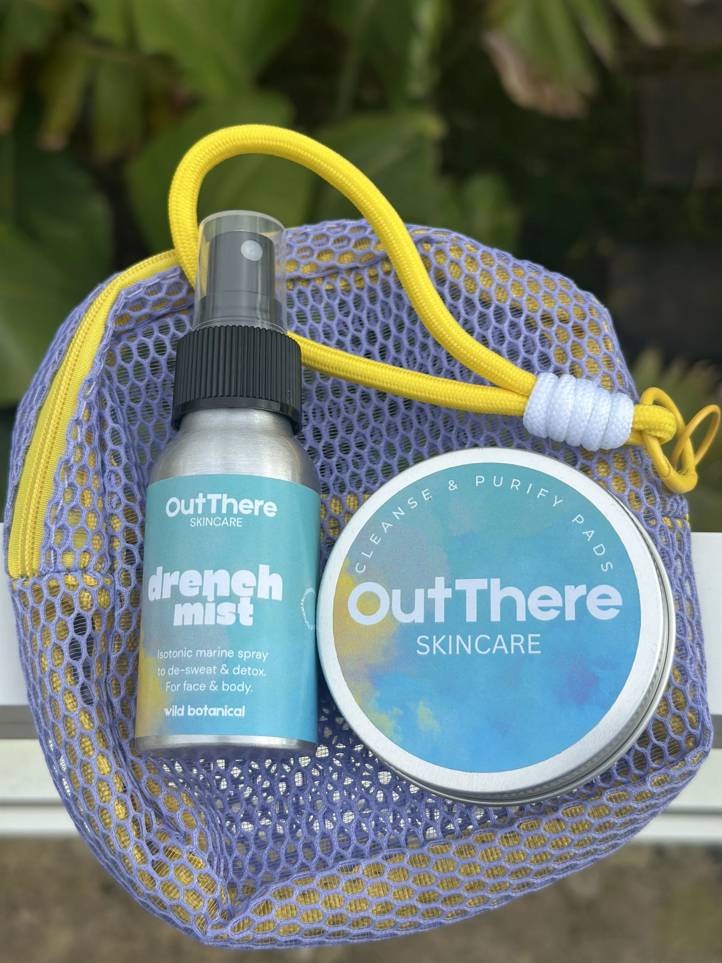 A purple mesh bag with a yellow strap contains a spray bottle labeled "drench mist" and a round container labeled "OutThere Skincare," being held outdoors with blurred green foliage in the background.