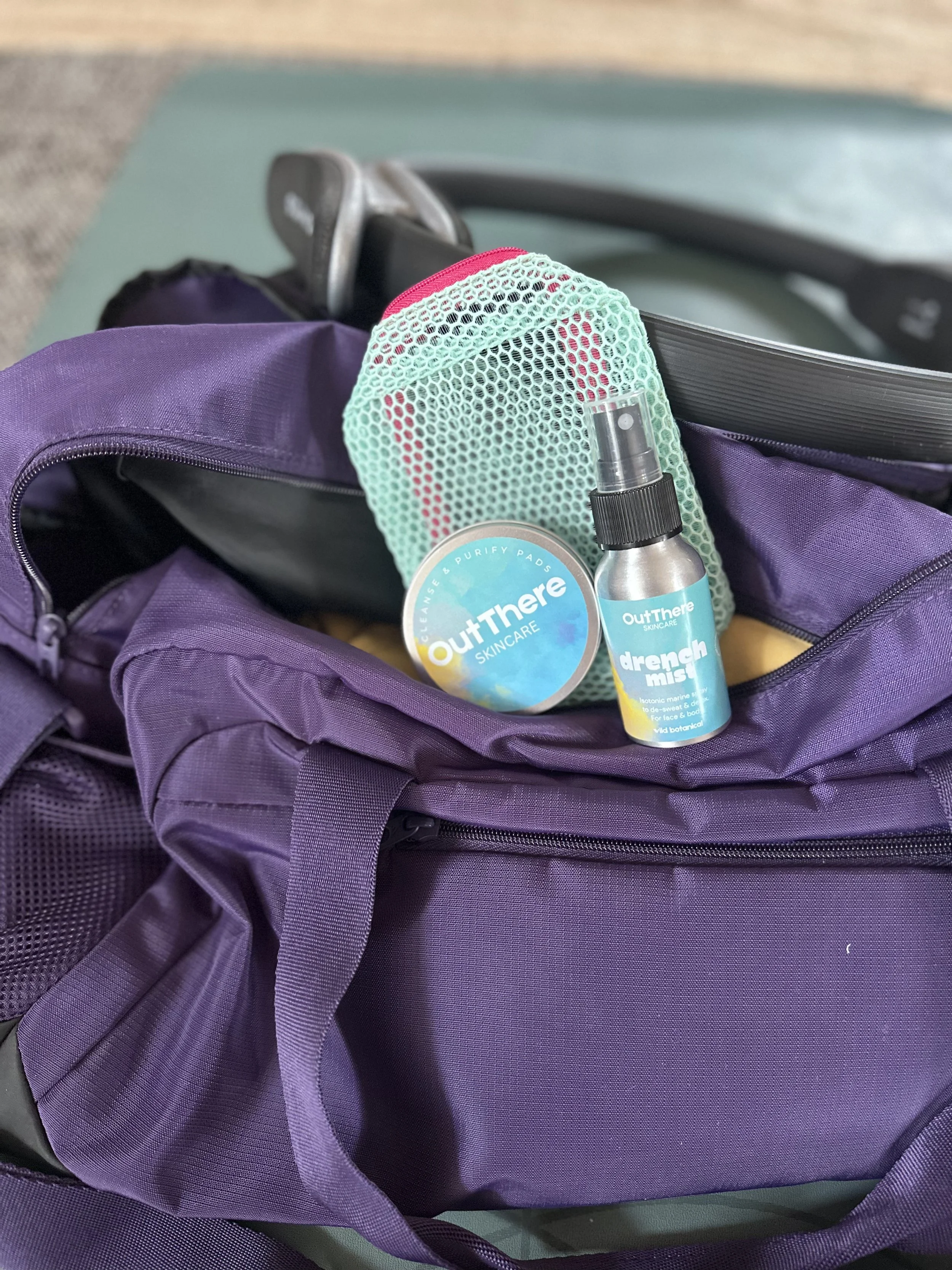 Open purple gym bag containing OutThere skincare products, including a seaweed and purify pads container, a Dreng mist spray bottle, and face and body wipes, with a tailgating chair in the background.