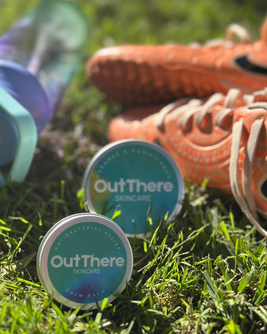 Outdoor scene with skincare products from outThere, a pair of orange sneakers, and a baseball glove laid on green grass.