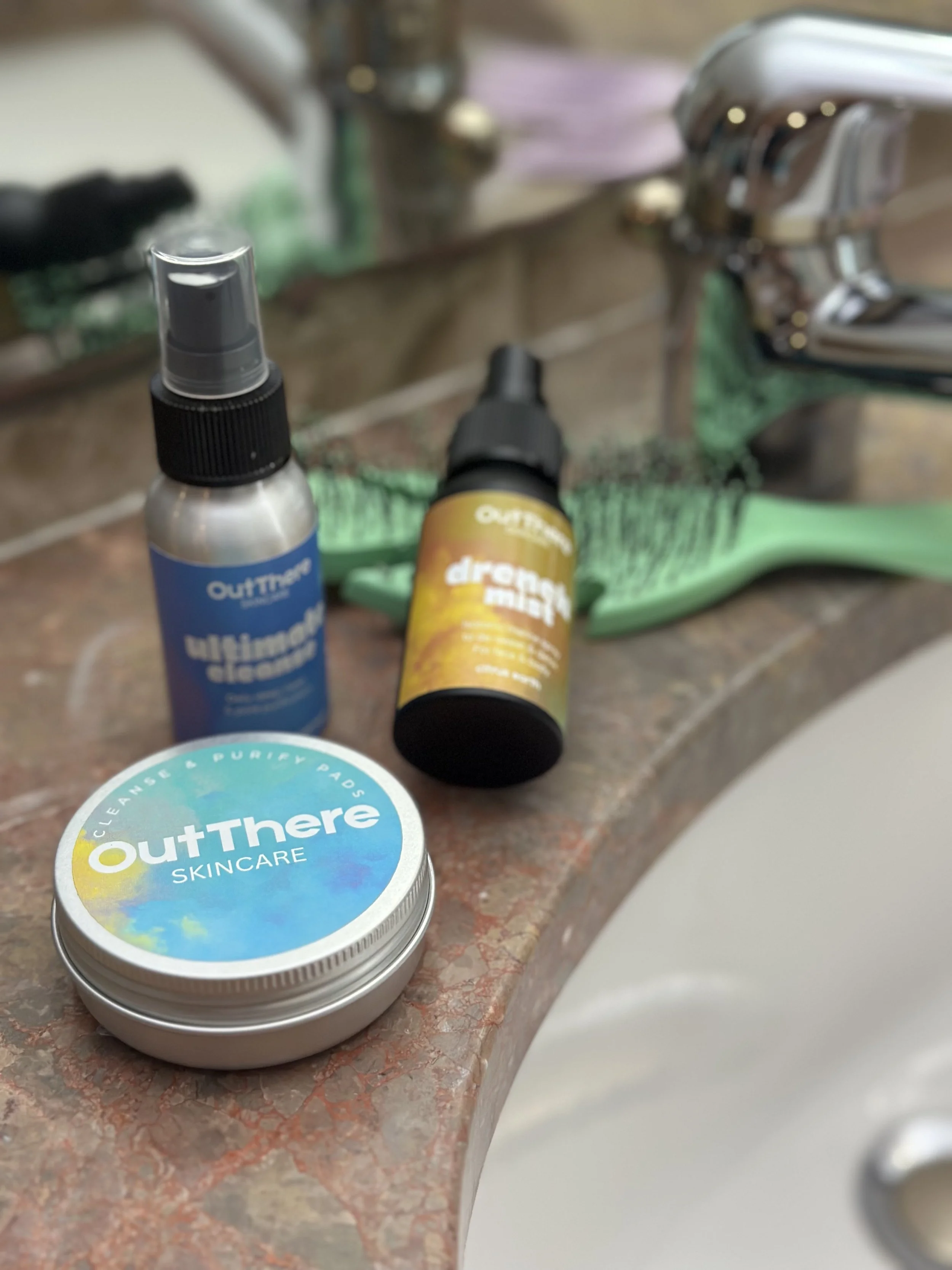 Skincare and grooming products on a bathroom sink countertop, including OutThere skincare, ultimate cleanser, and a mister, with a green toothbrush in the background.