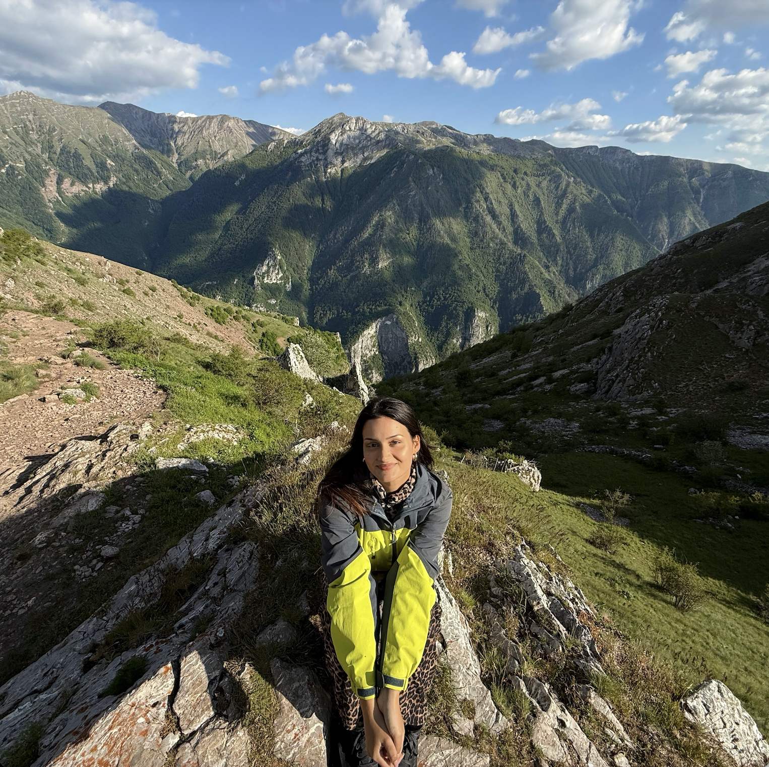 A woman with dark hair, wearing a gray and yellow jacket, is sitting on a large rock in a mountainous landscape with green hills, rocky terrain, and mountain peaks in the background under a partly cloudy sky.