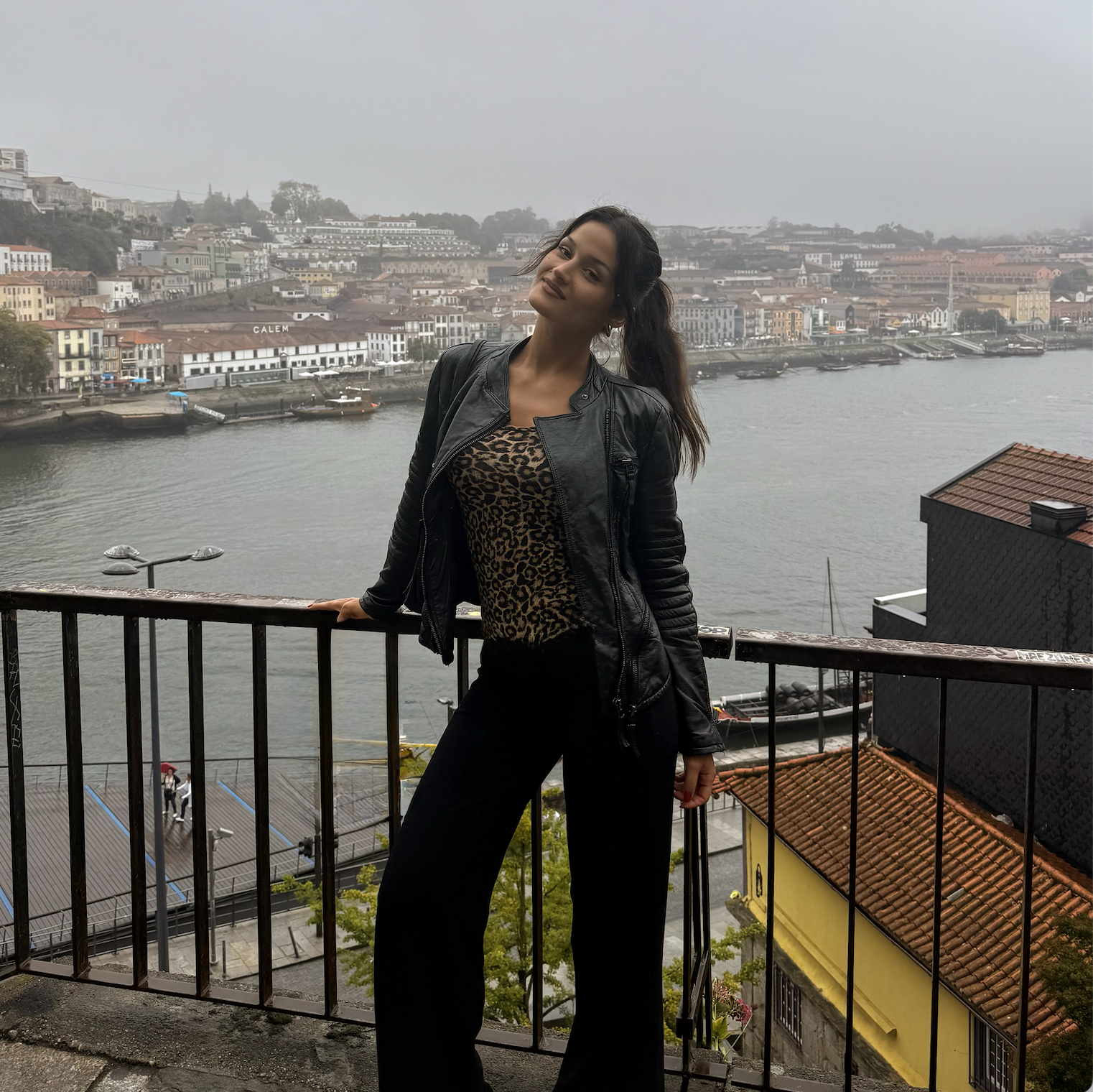 A woman with long dark hair standing on a balcony overlooking a river with buildings on the opposite bank. She is wearing a black leather jacket and leopard print top, with one hand resting on the railing and the other holding a small bag. The weather is overcast and misty.