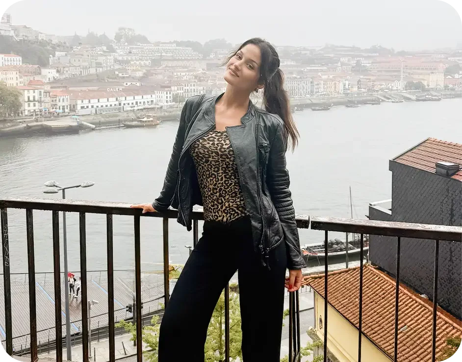 Young woman standing on a balcony overlooking a river with buildings and boats in the background, wearing a leather jacket and leopard print top.