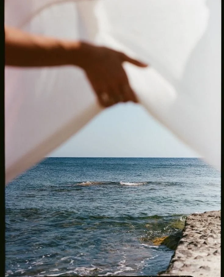 A view of the ocean through a shaped opening made by a person's hands and white fabric, with rocks in the foreground and gentle waves on the water.