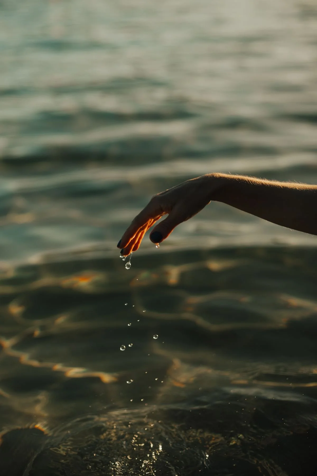 A hand with black painted nails is dipping into water, with water droplets falling from the fingers, over a body of water at sunset.