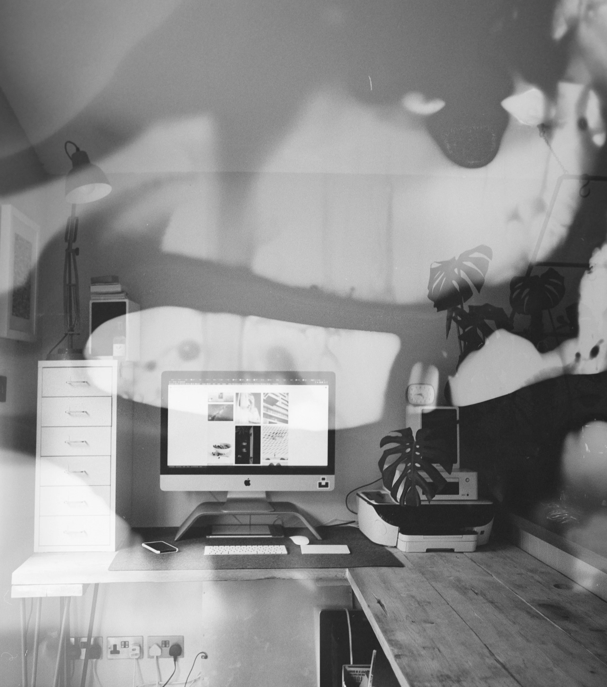 Photo d'un bureau avec un ordinateur iMac, une petite caisse de rangement, une imprimante, et des plantes d'intérieur, avec une lumière tamisée et des éléments décoratifs sur le mur.