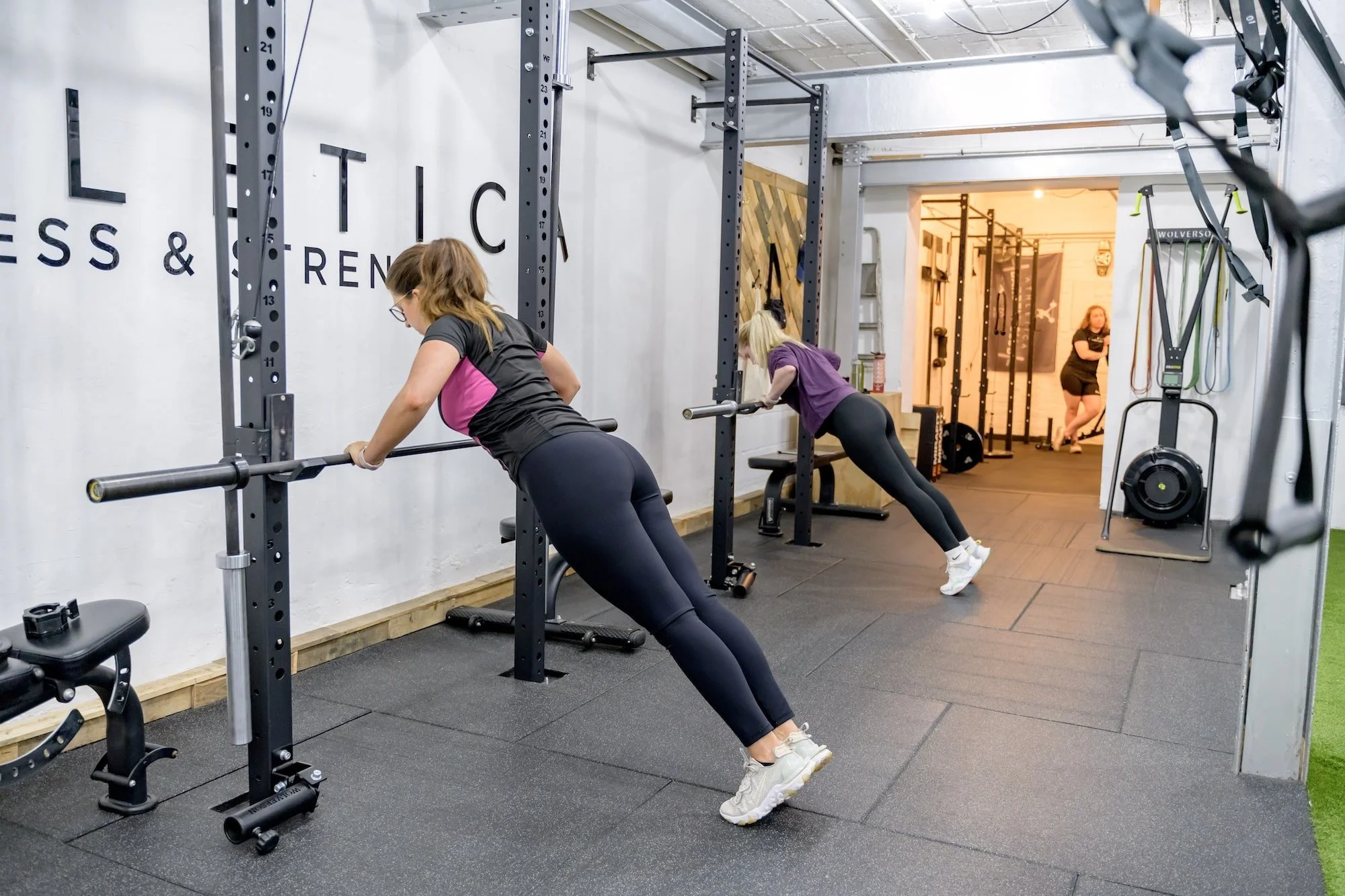 Two women doing inclined push-ups using barbells in a gym with black rubber flooring, weight racks, and fitness equipment.