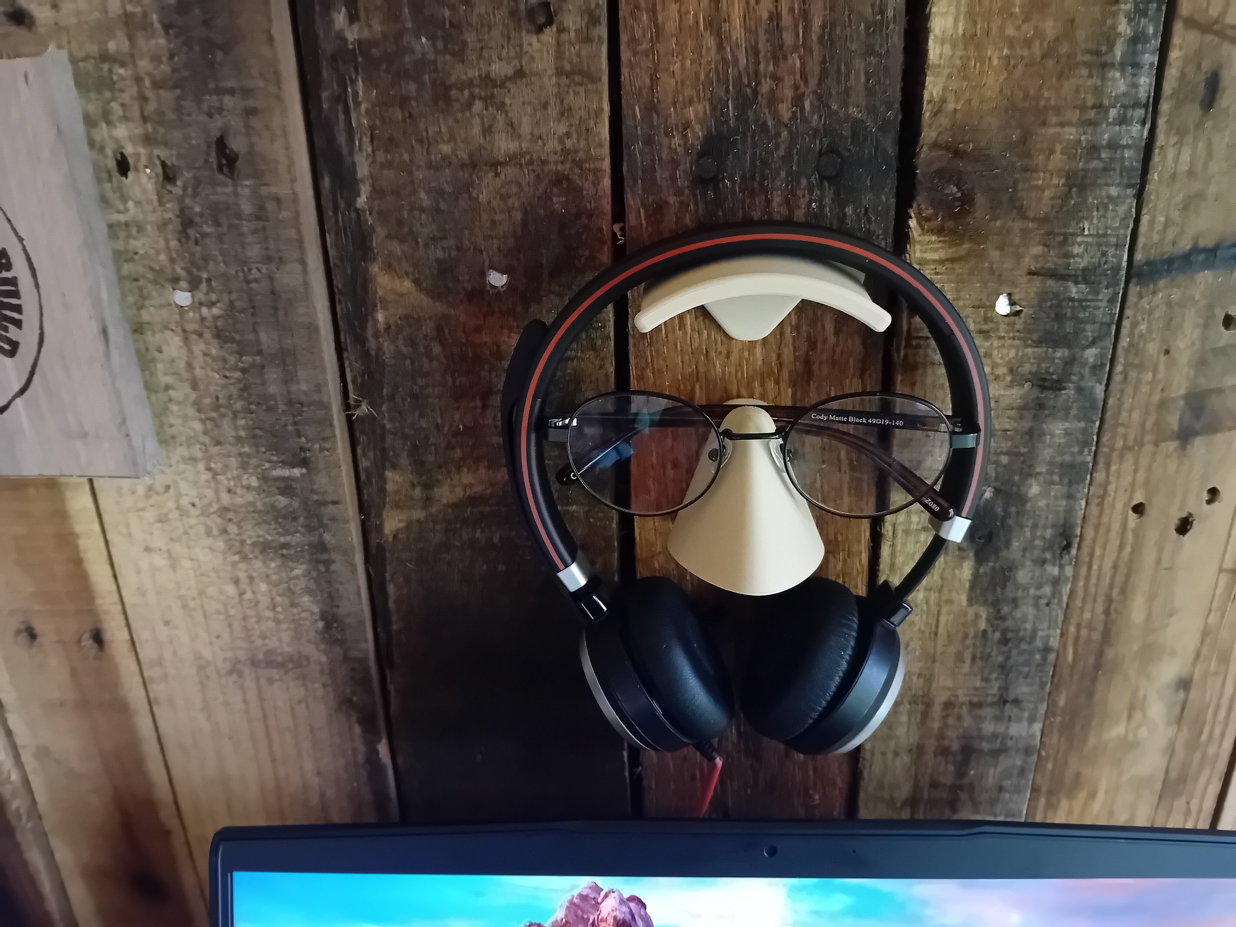 A pair of eyeglasses resting on a beige nose-shaped stand, with over-ear headphones hanging around the stand, all placed on a rustic wooden surface.