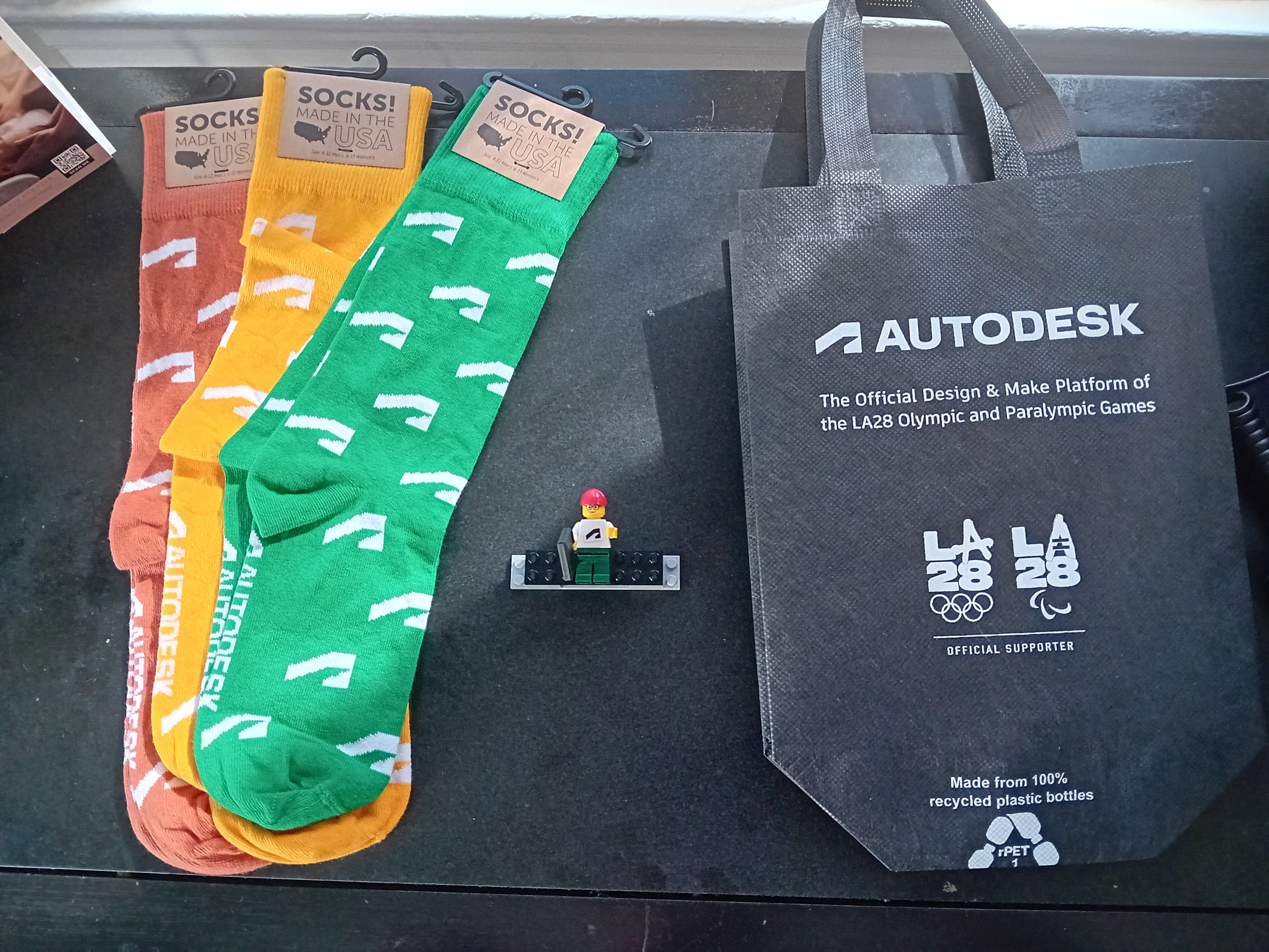 A set of three pairs of colorful socks with repeating logo patterns, a LEGO minifigure holding a skateboard, and an Autodesk branded tote bag on a black surface.