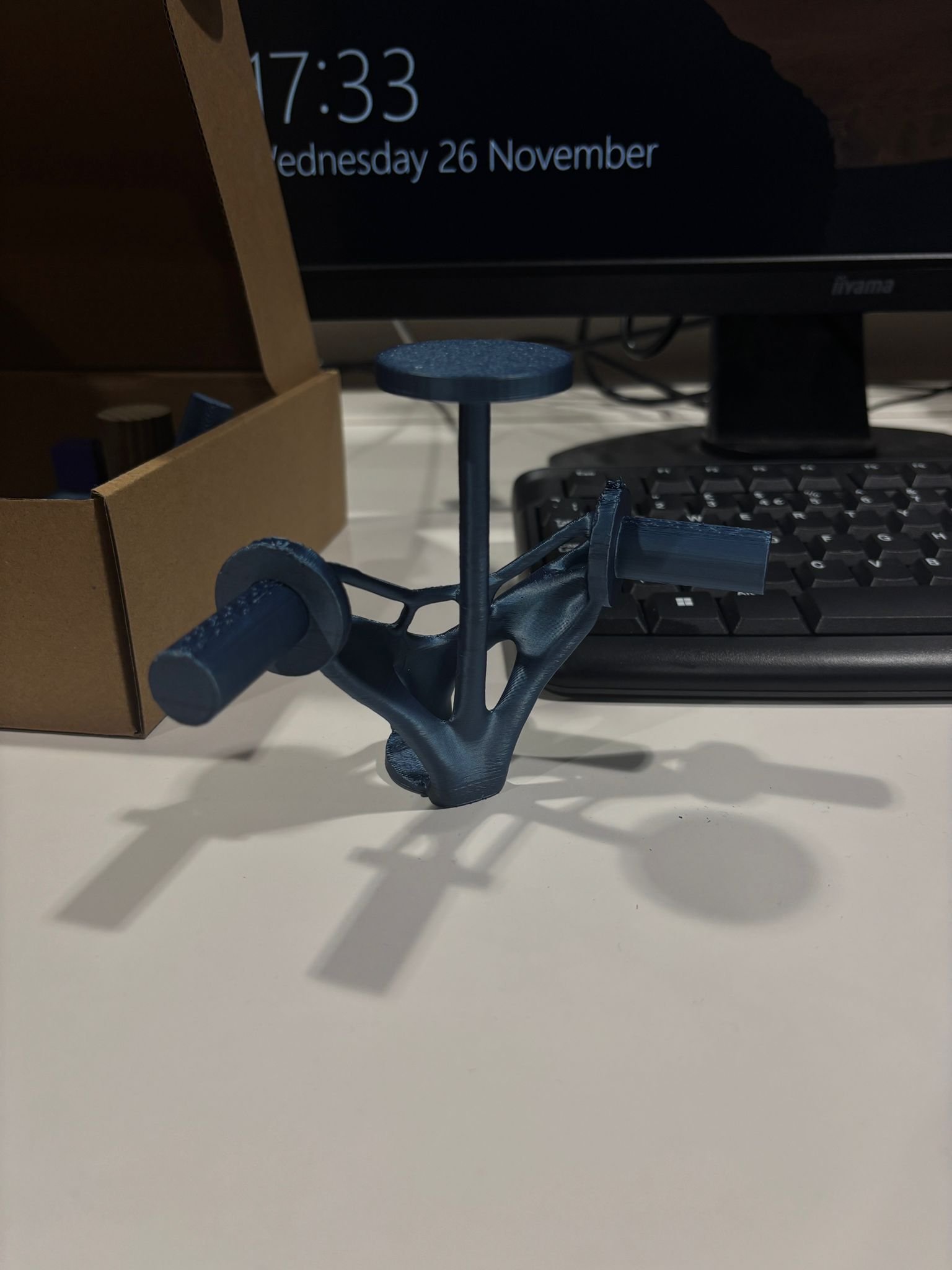 3D printed black part with cylindrical extensions, casting a shadow on a white surface in front of a computer monitor, keyboard, and cardboard box.