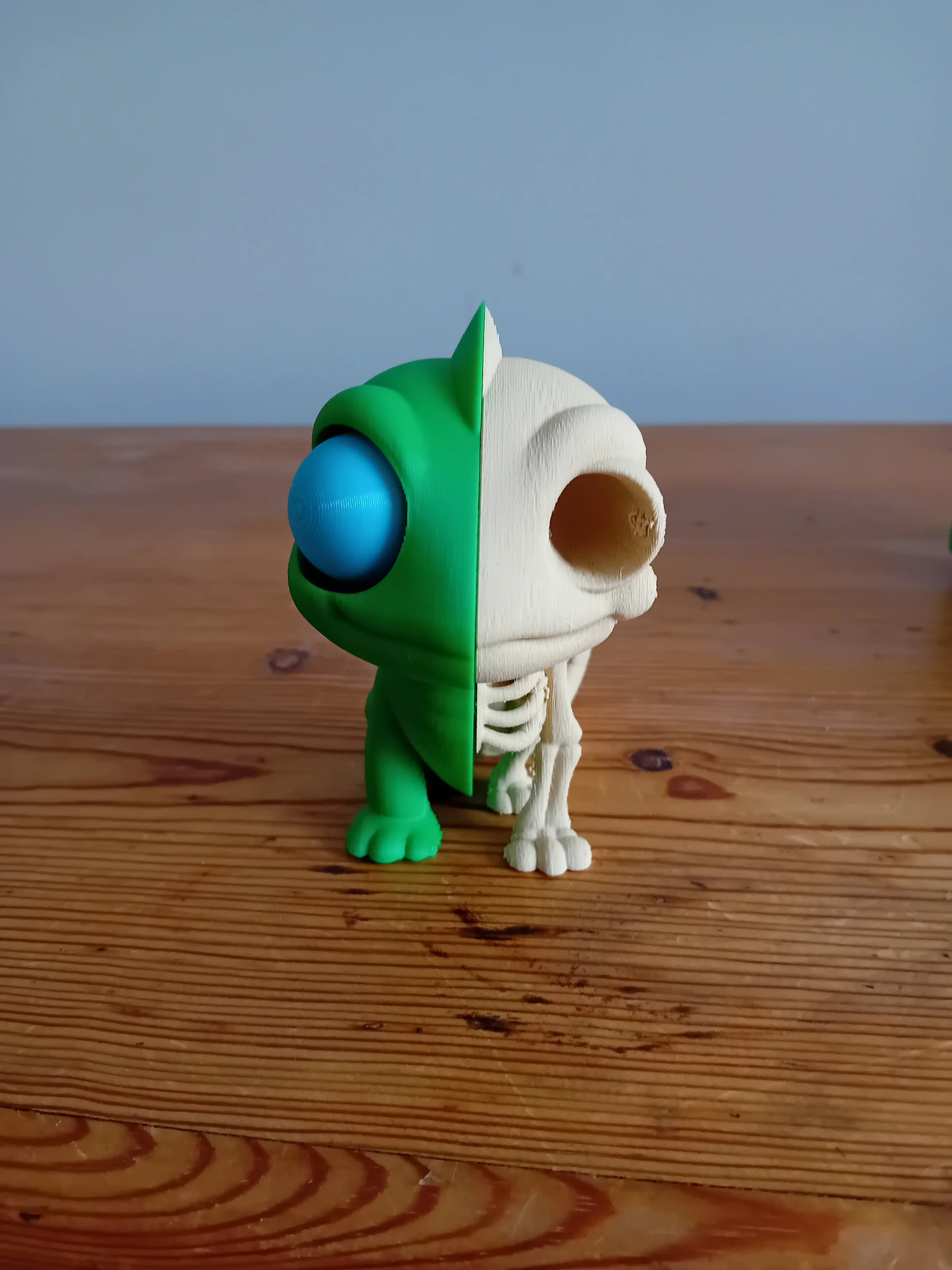 Half of a green cartoon robot with a blue eye and a helmet, and half a white skeletal dog model, arranged in a single figure, placed on a wooden surface.