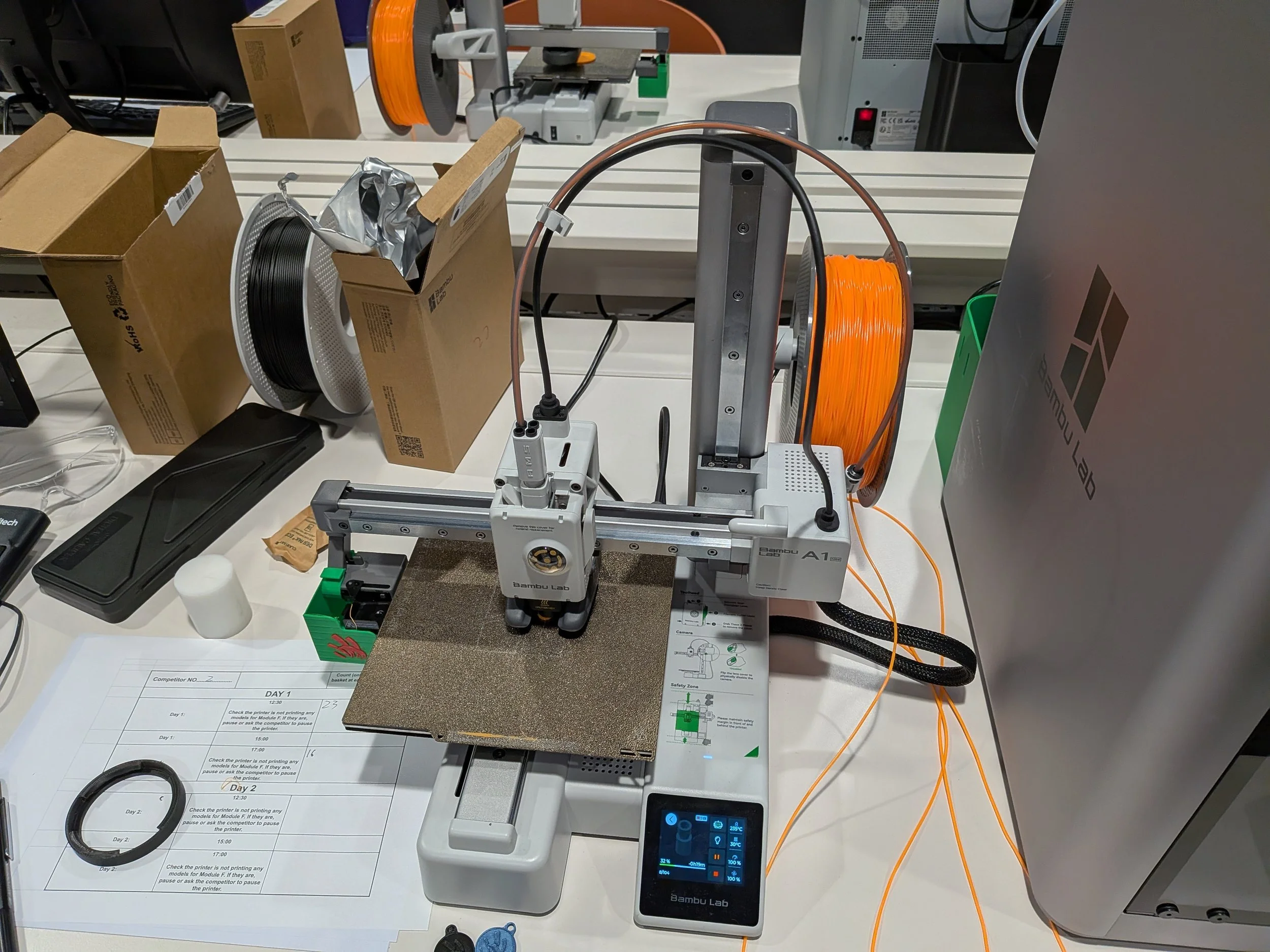 A desktop workspace with 3D printing equipment, including a white Bambu Lab 3D printer with orange filament spool, cardboard boxes, a computer mouse, a small control panel with a blue screen, and various technical components.