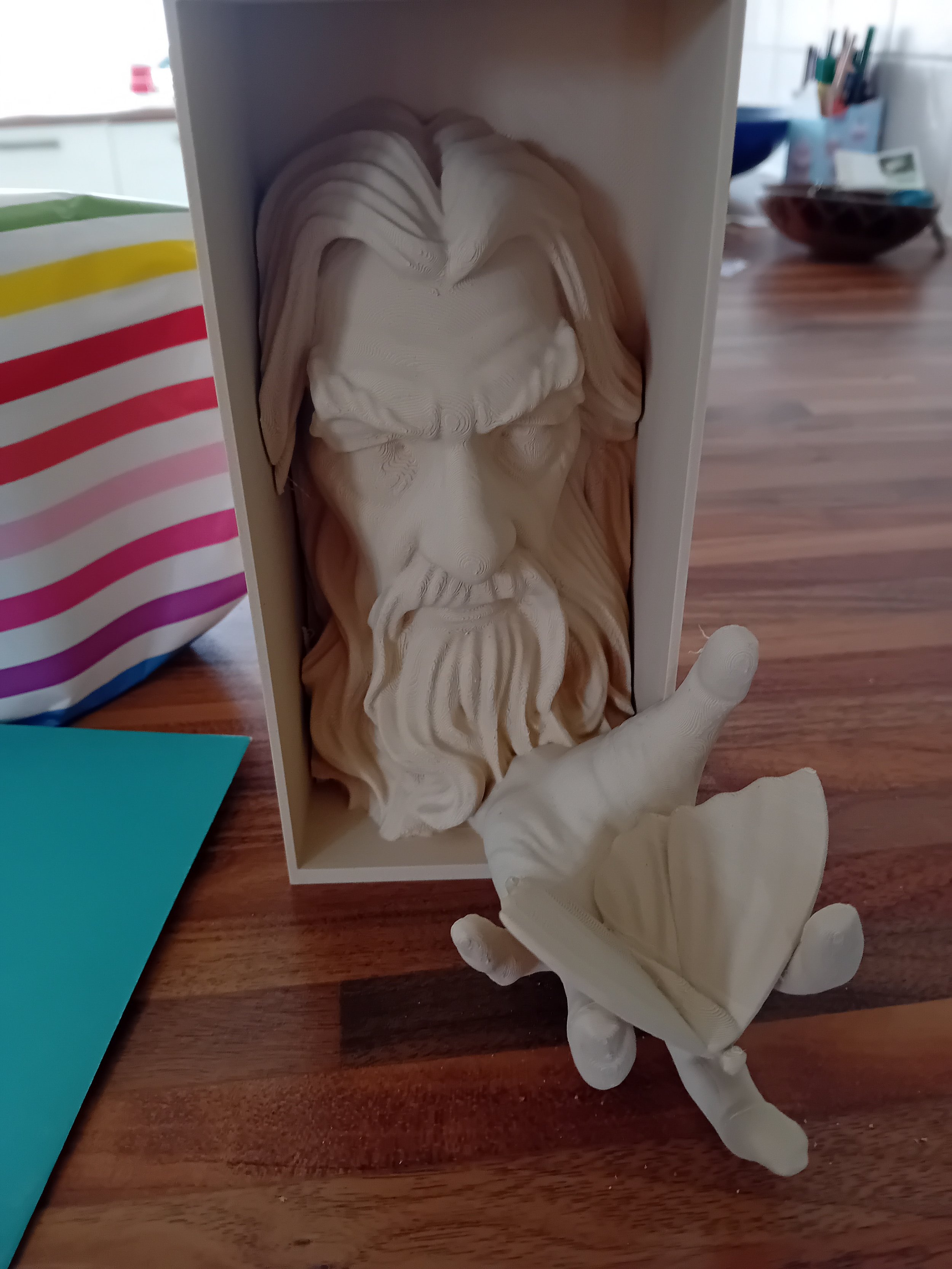 A white sculpture of a bearded man's face with long hair, inside a box on a wooden table. An arm and hand are attached to the sculpture, extending outward with the hand open.