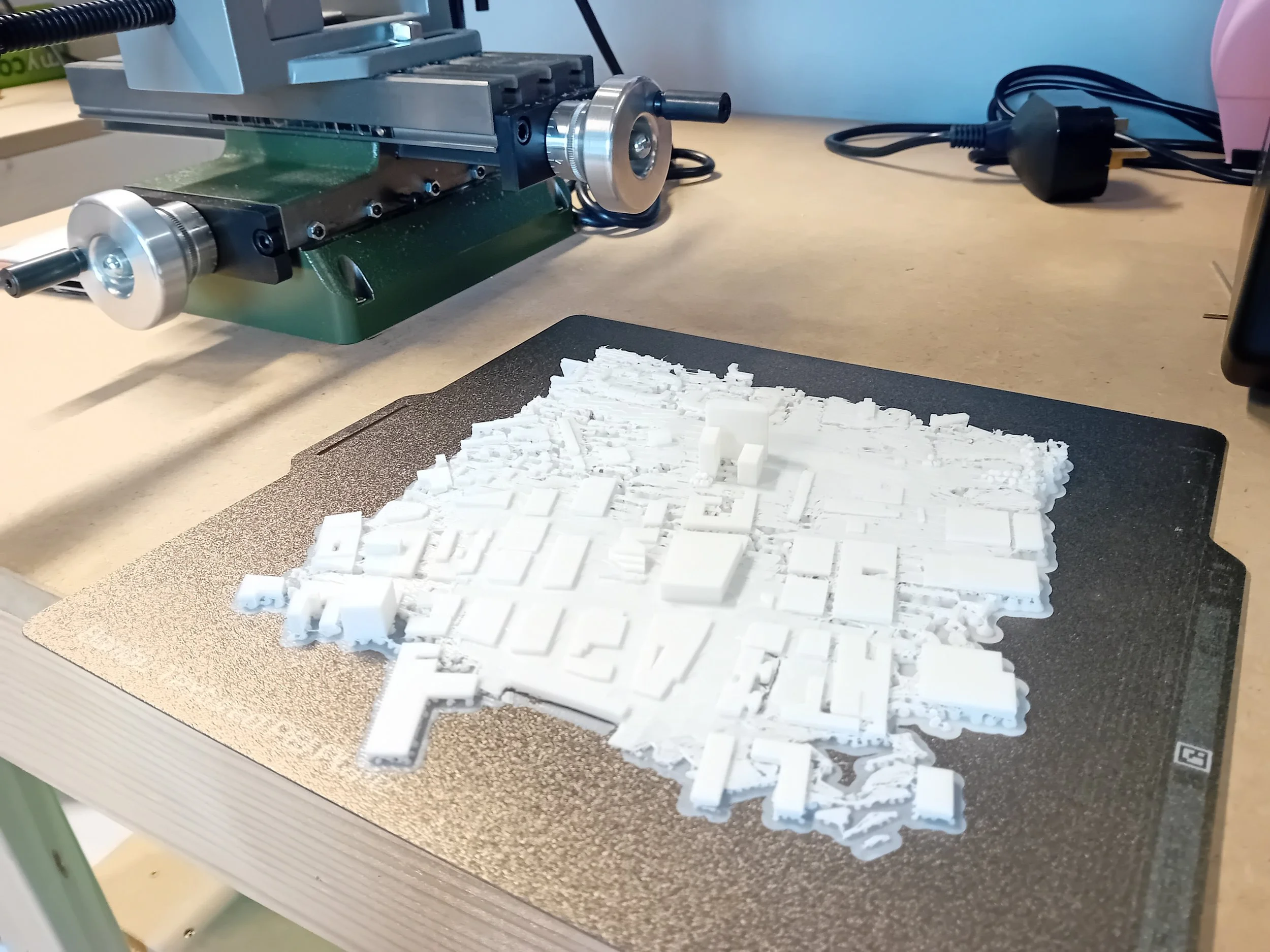White 3D-printed city model on a black platform, with a mechanical device nearby on a workbench.
