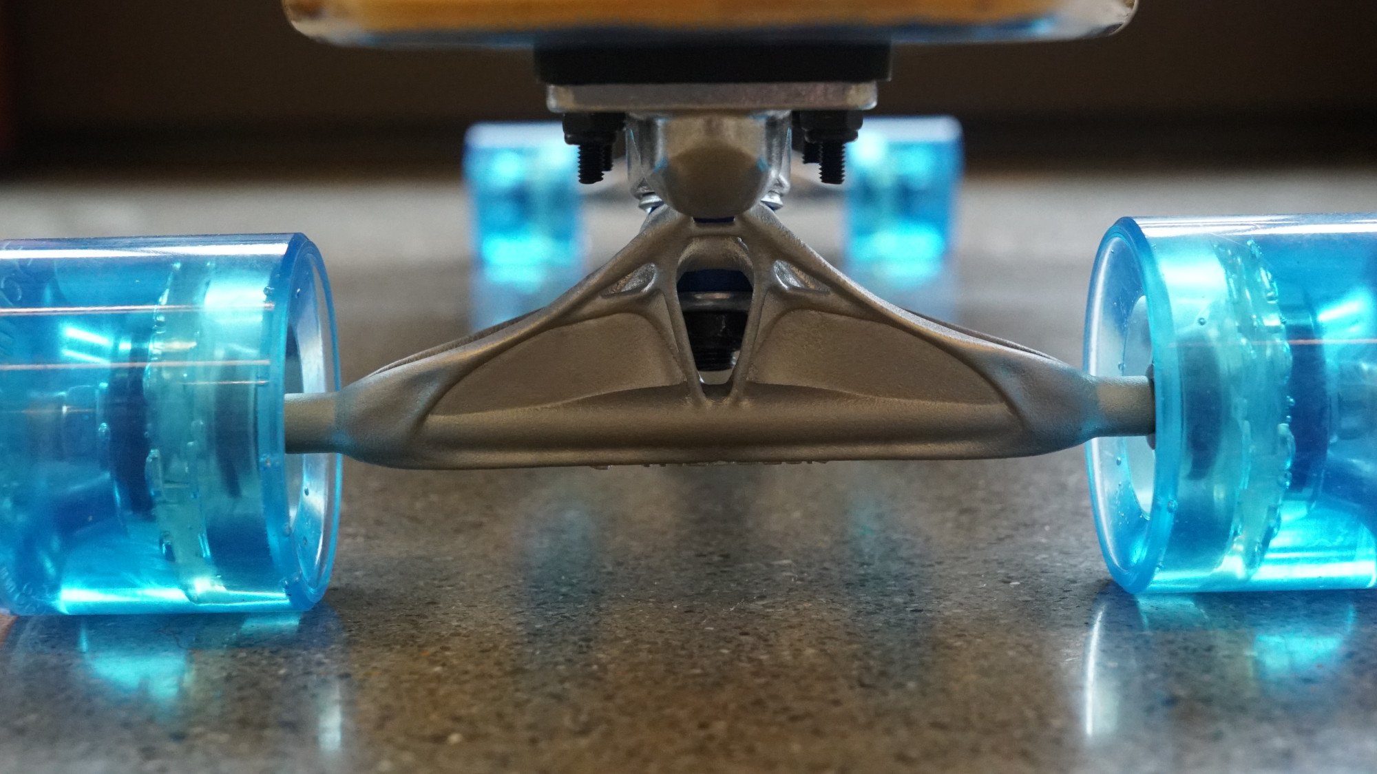 Close-up of a skateboard wheel assembly, showing two translucent blue wheels attached to a metal truck on a textured surface.