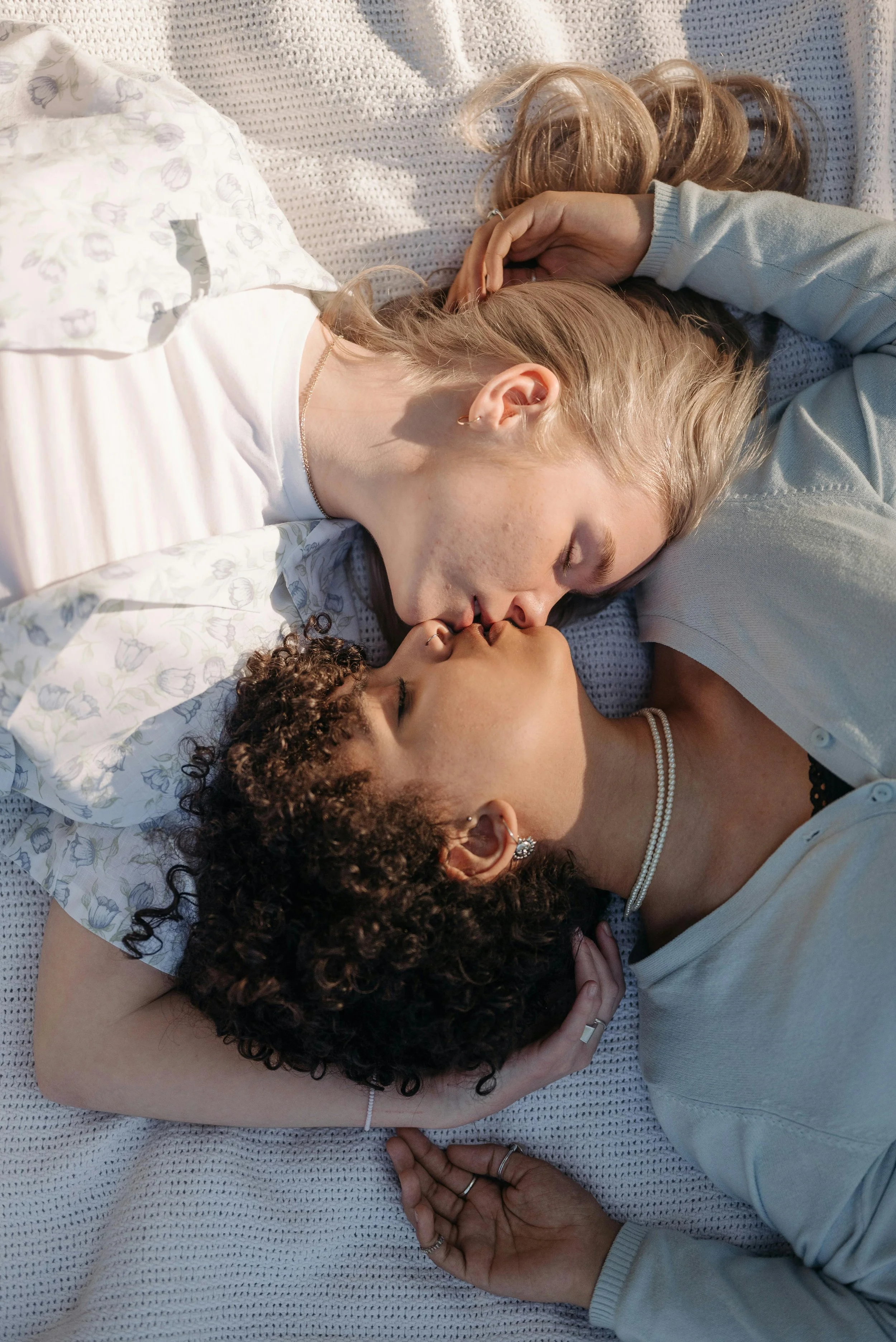 Two women with different skin tones and curly hair lying on a bed, kissing each other.