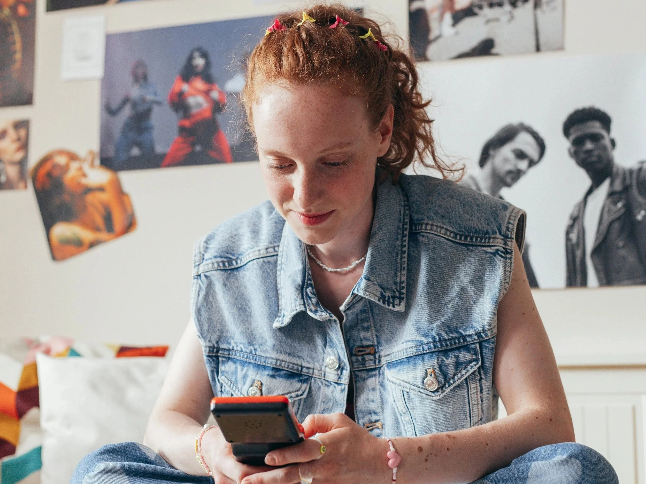 A young woman with red curly hair, wearing a denim vest and jewelry, is sitting on a bed and looking at her phone. The background features wall art with images of people.