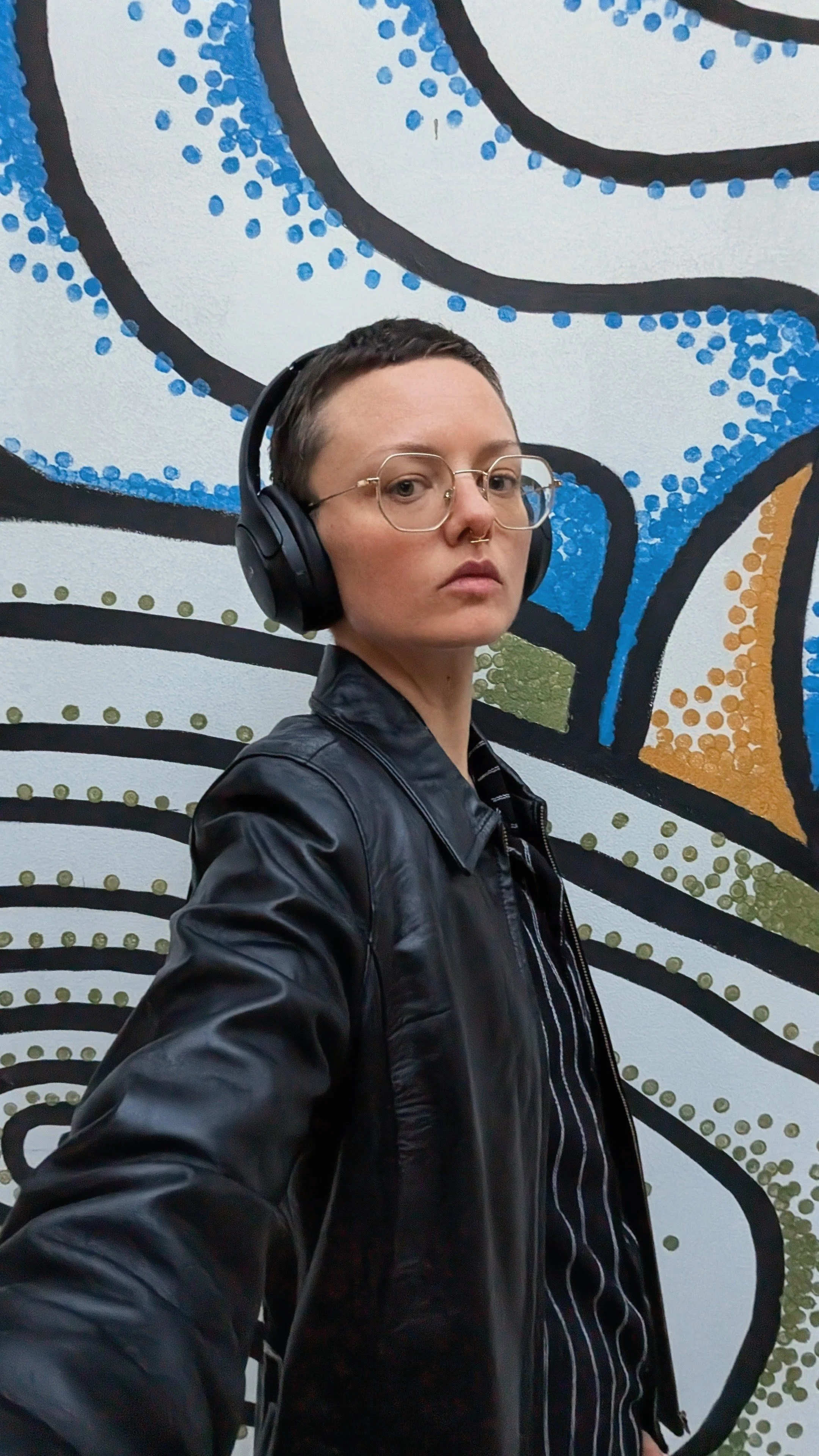 A person with short hair, glasses, and a septum piercing wearing large black headphones and a black leather jacket stands against a colorful, abstract mural background.