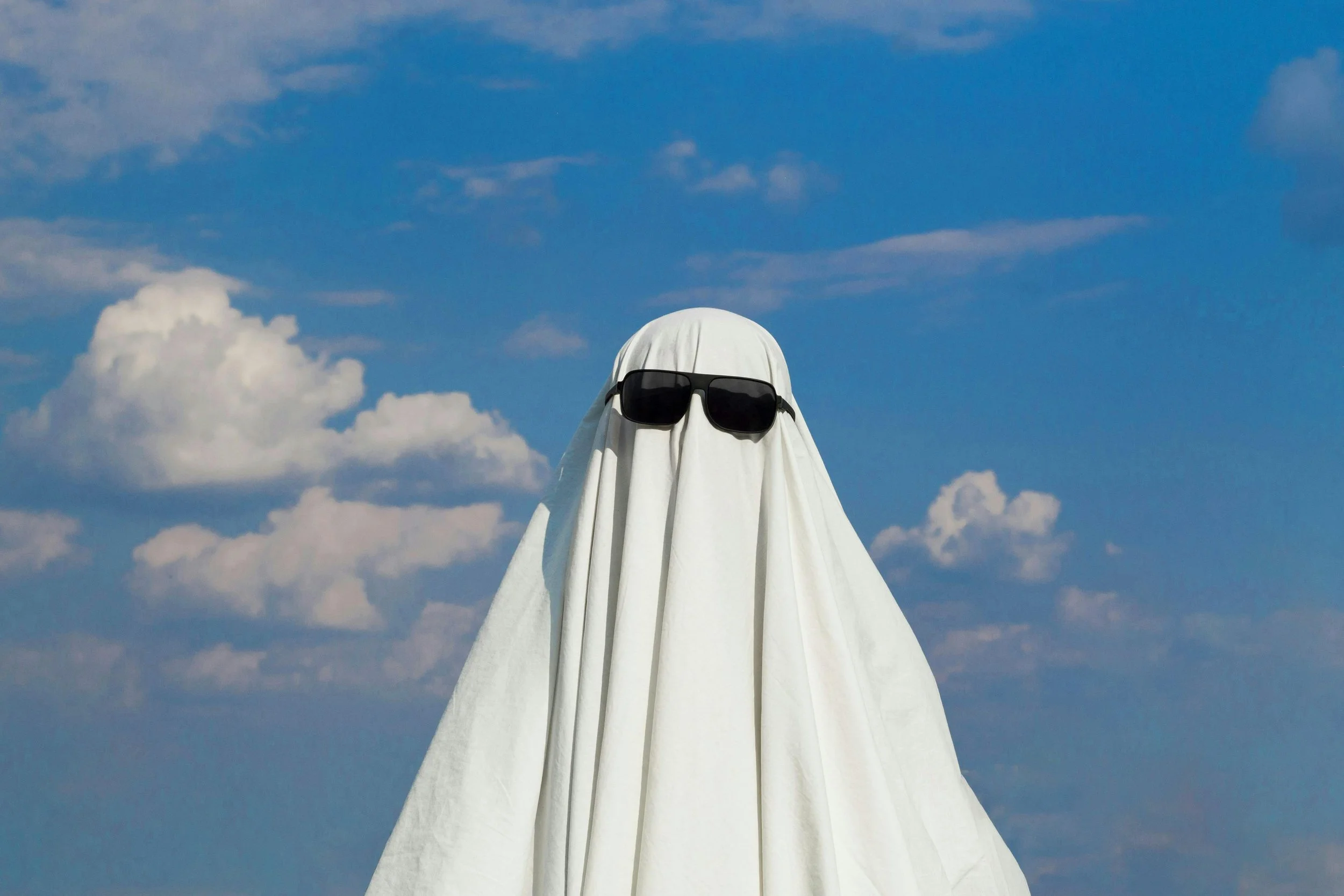 A person dressed as a ghost with white fabric and glasses, standing outdoors under a blue sky with clouds.