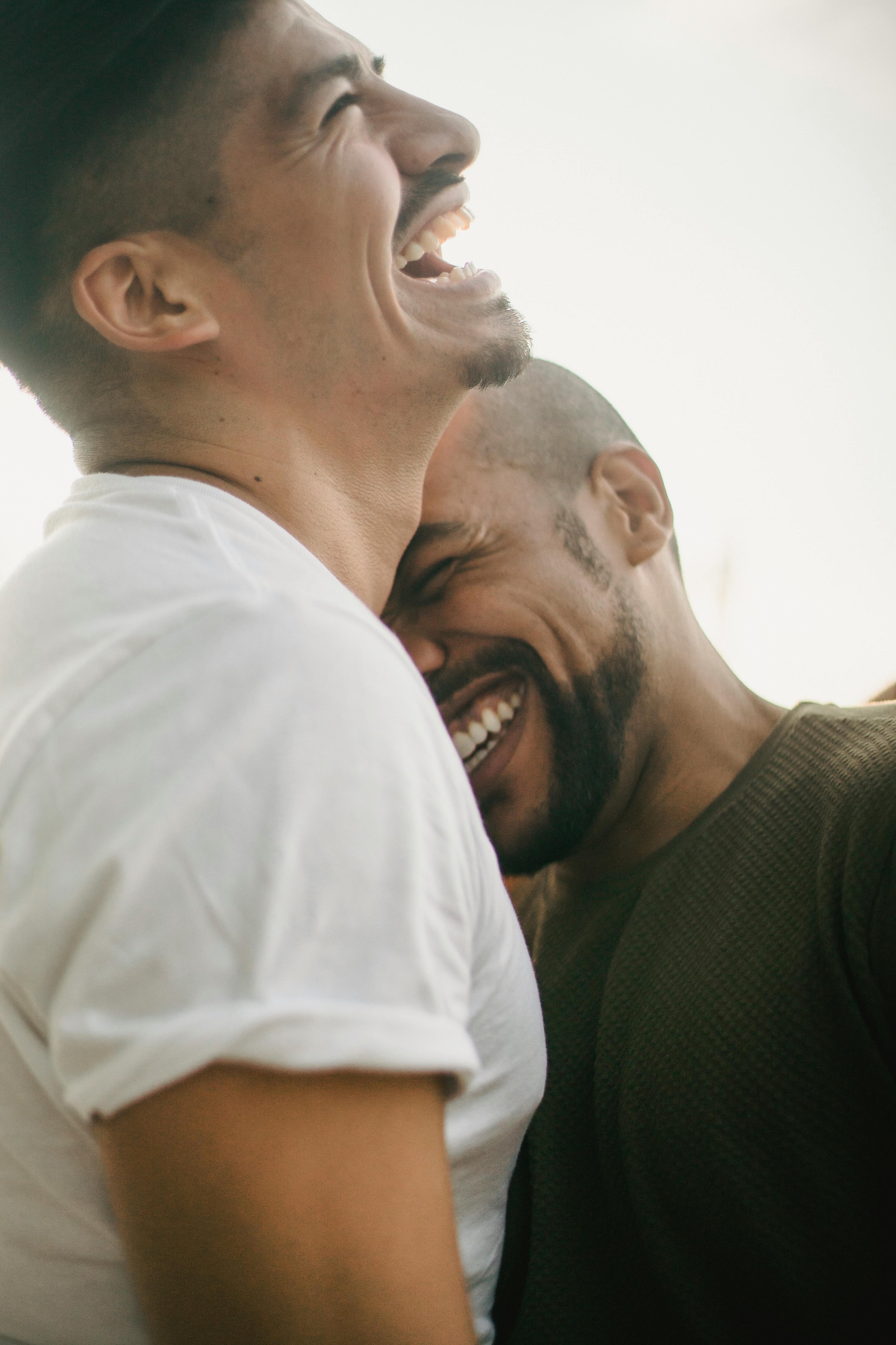 Two men are laughing and hugging each other, one is wearing a white t-shirt and the other is in a dark shirt.
