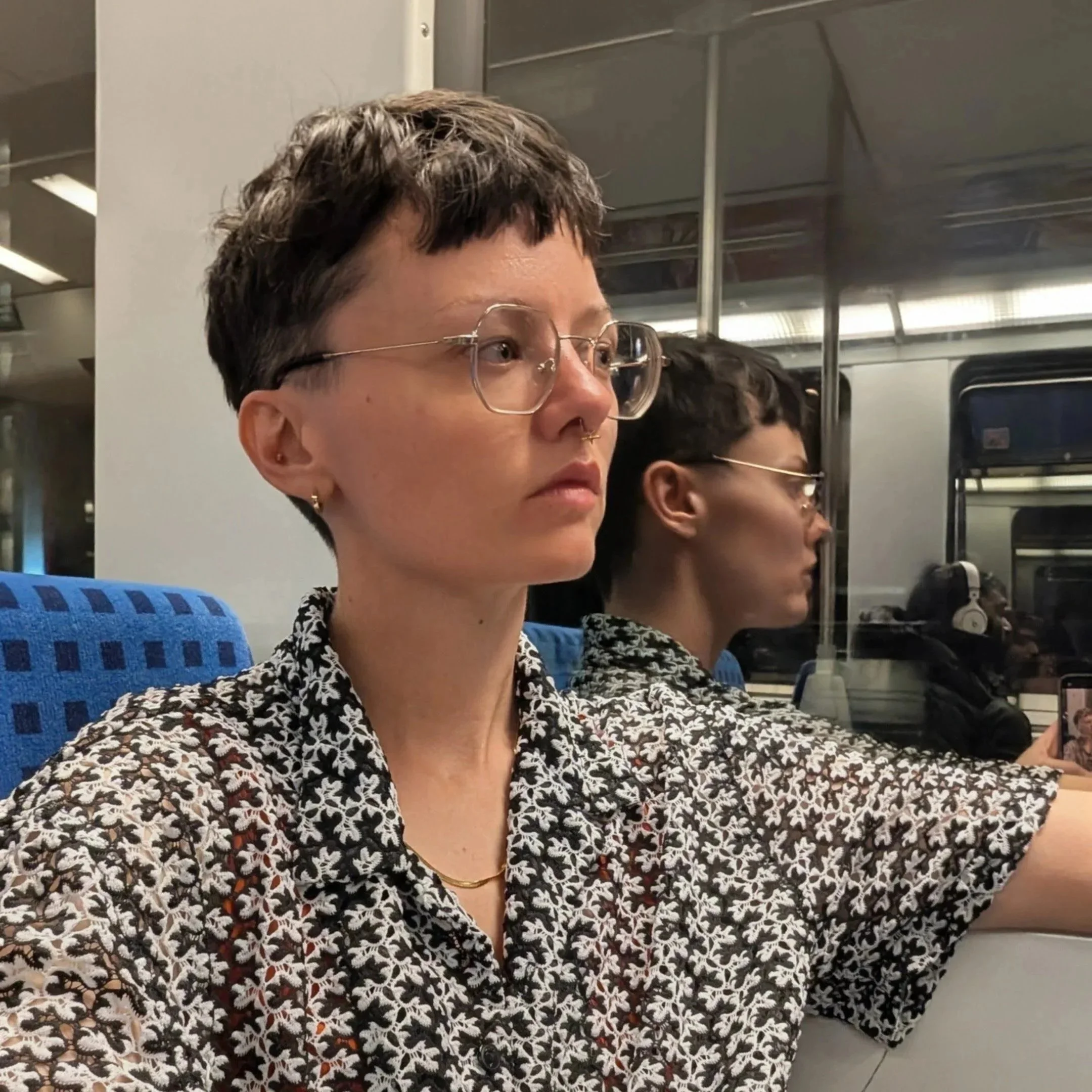 A person with short dark hair, glasses, and a patterned shirt sitting on a train, looking straight ahead. Their reflection is visible in the window behind them.