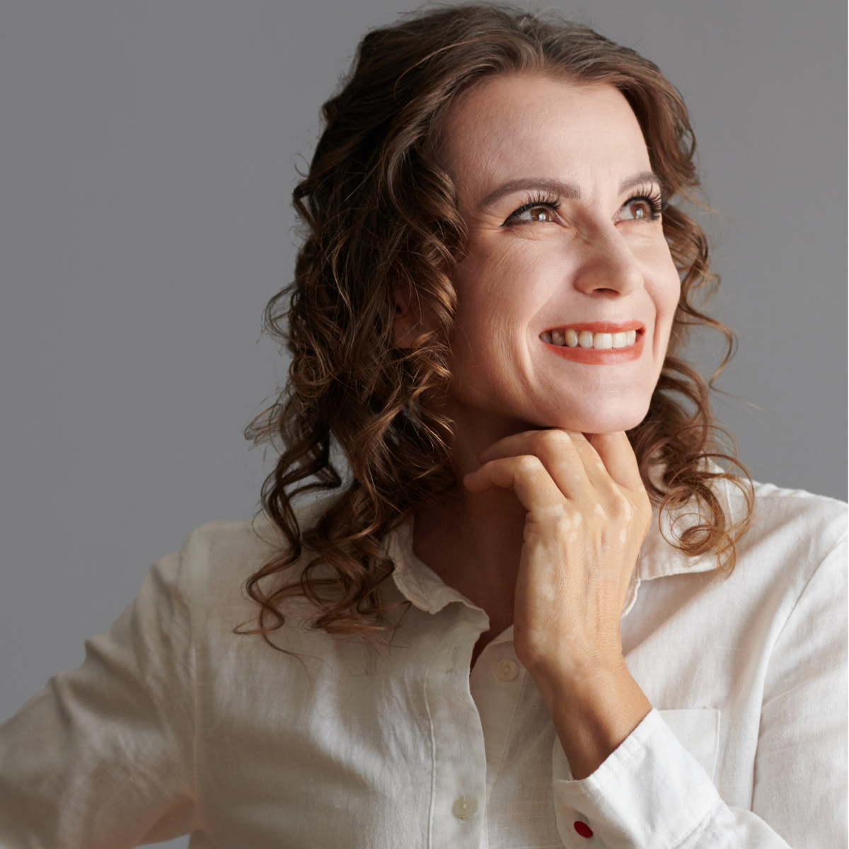 Portrait of a smiling woman with curly brown hair, wearing a white shirt, looking upward with her right hand resting on her chin.