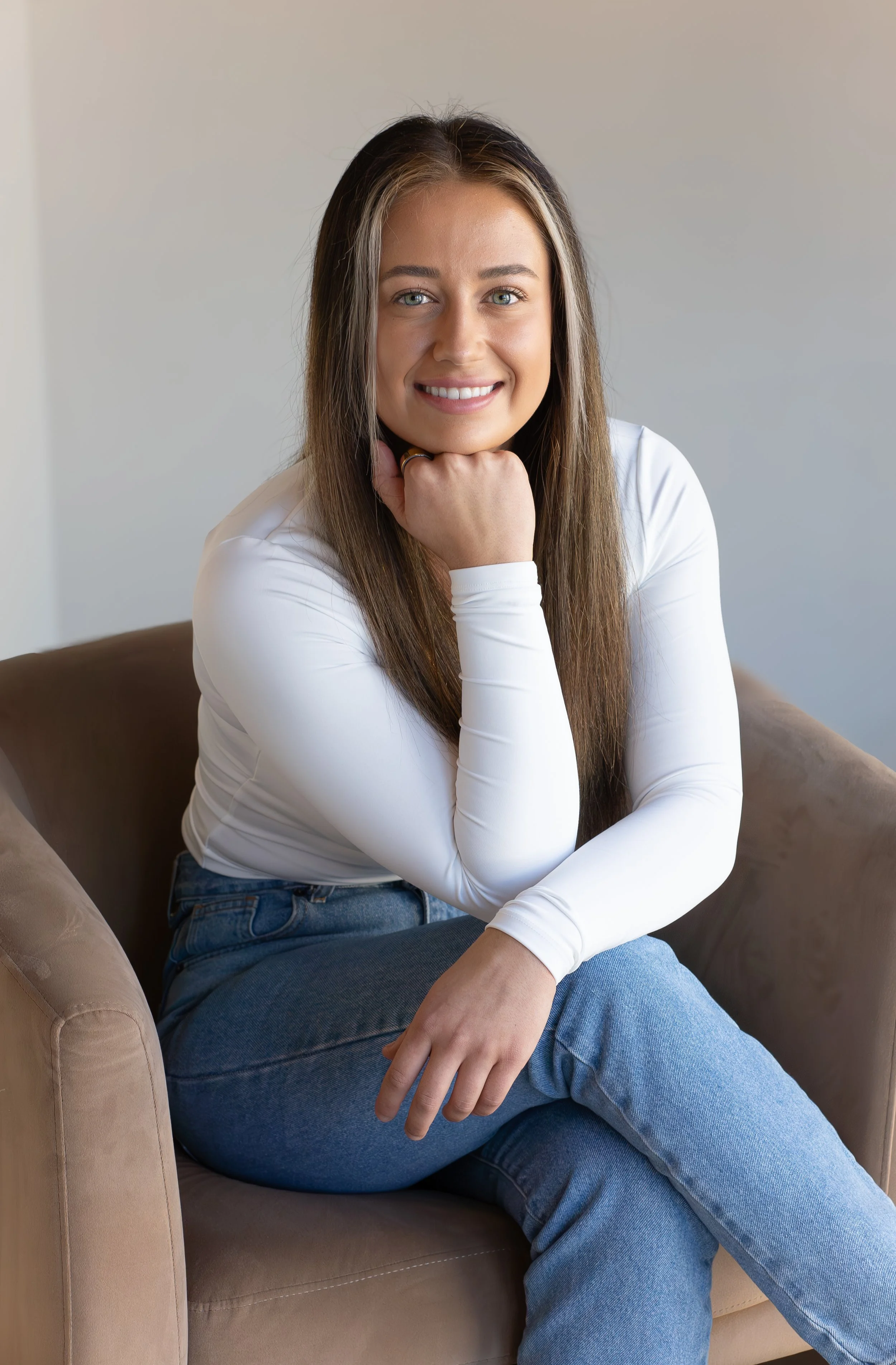 A woman with long brown hair and blue eyes sitting on a brown armchair, smiling, wearing a white long-sleeve shirt and blue jeans, with her chin resting on her hand.