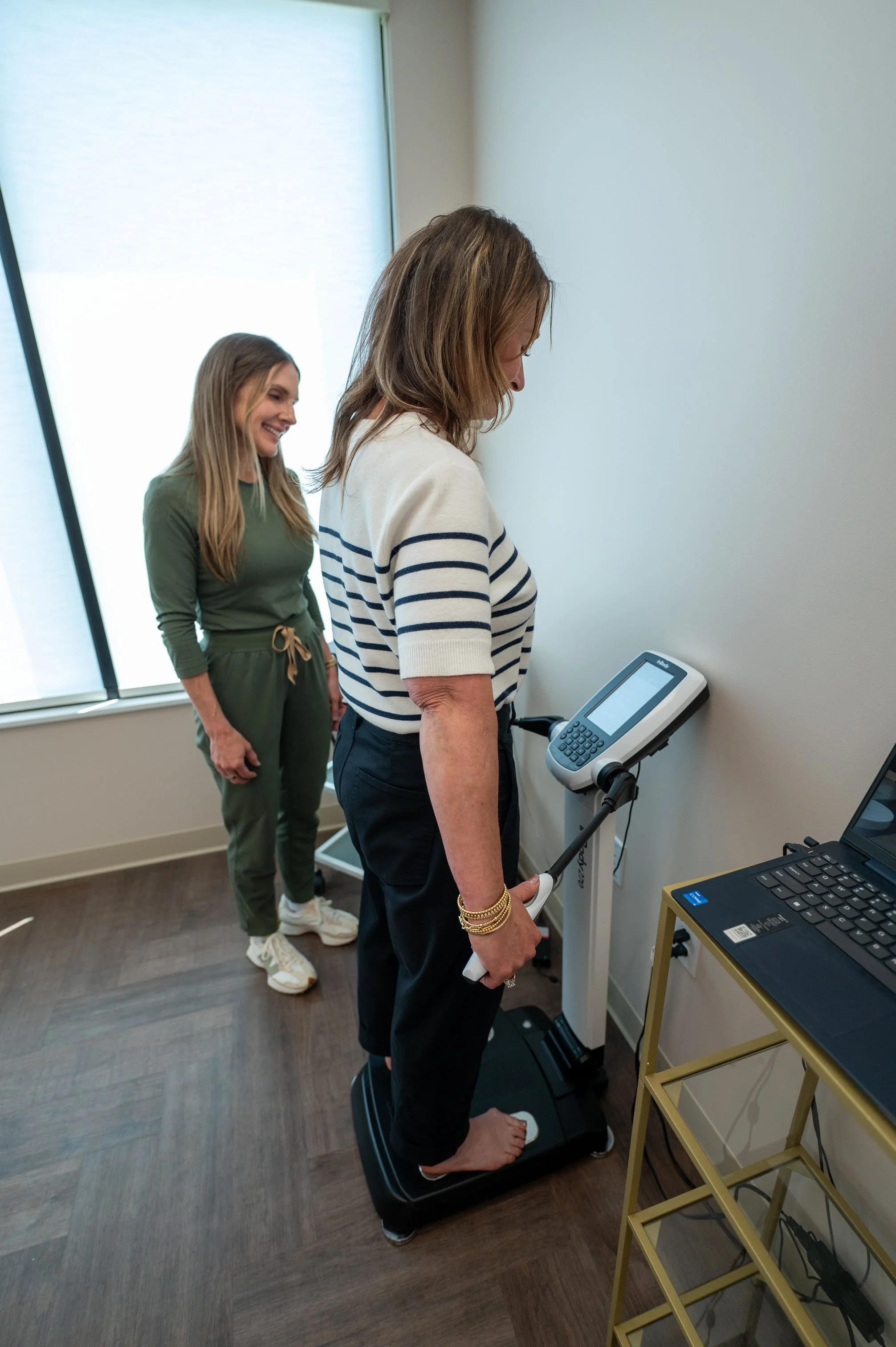 Two women, one older and one younger, are using a treadmill with a built-in display in a room with wooden flooring and a large window with blinds. The younger woman is standing on the treadmill, holding a device, and looking at the display, while the older woman is standing nearby and smiling. A yellow shelving unit with electronic devices and a keyboard is visible in the room.
