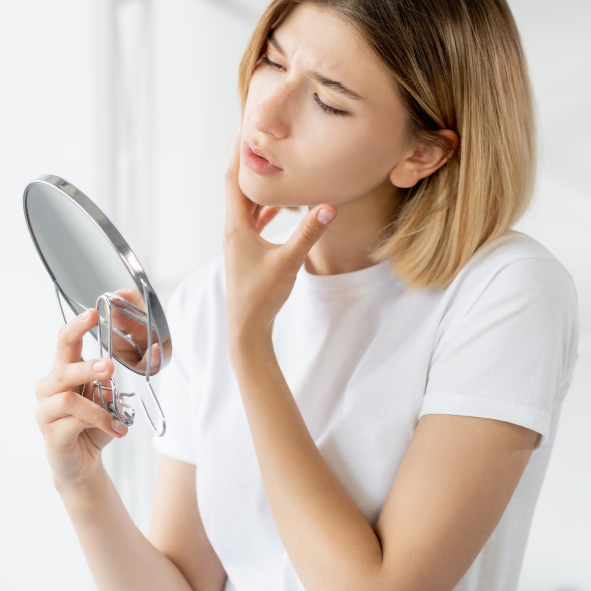 A young woman with shoulder-length blonde hair looks at herself in a small round mirror, with a confused or concerned expression, touching her chin with one hand.